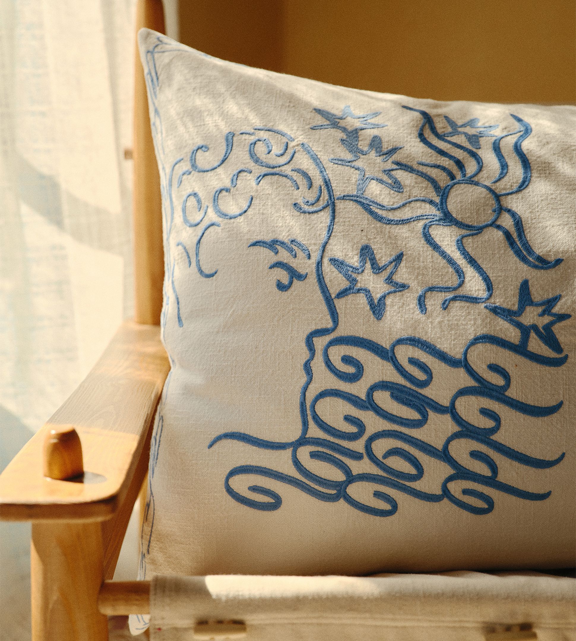 Un coussin beige sur une chaise en bois, orné d'une broderie bleue raffinée représentant une silhouette, des étoiles et un soleil. La lumière chaude du soleil filtre à travers un rideau.