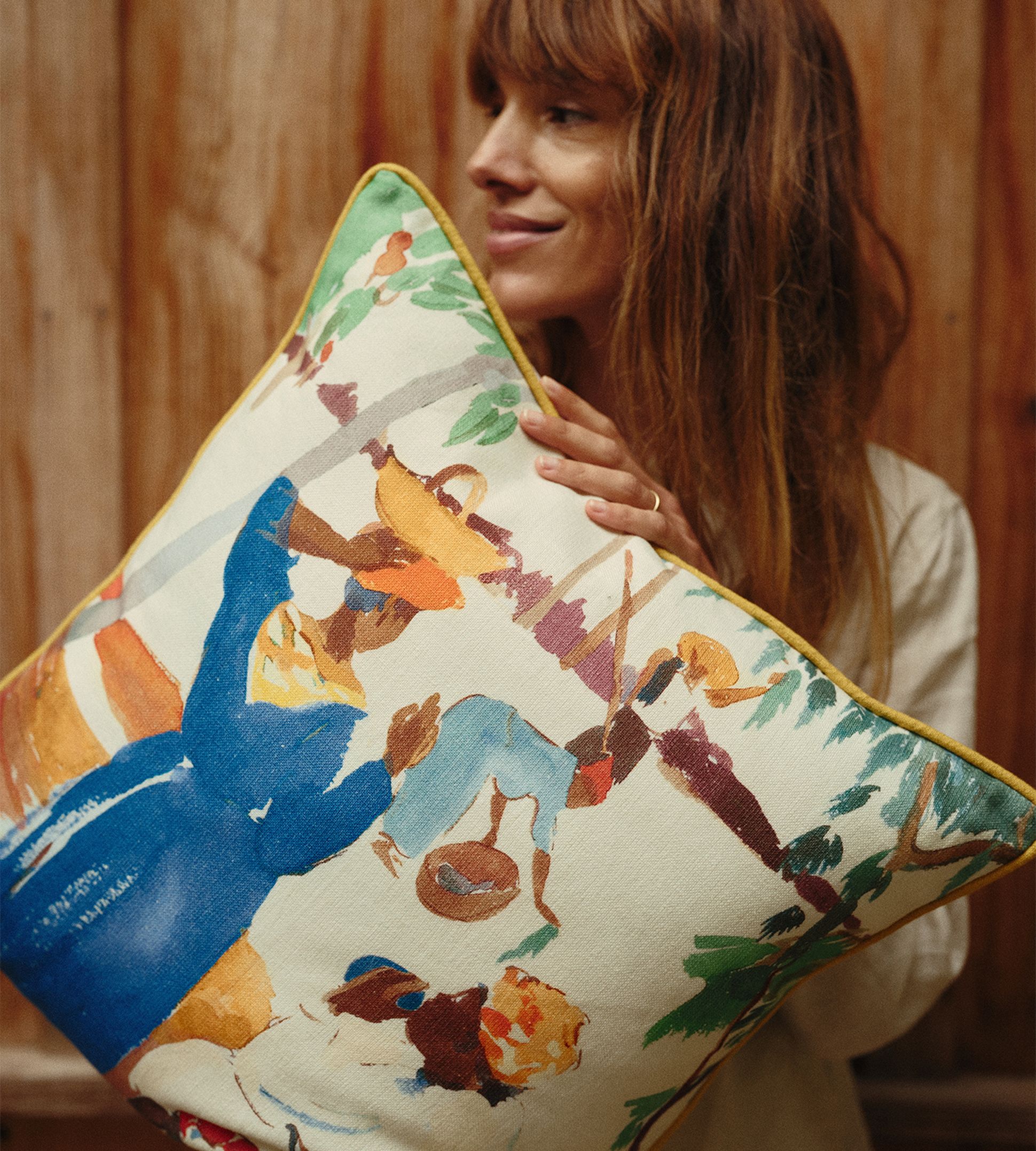 Une femme tenant un coussin coloré orné d'une peinture abstraite et vivante représentant des personnages, sur fond de bois.