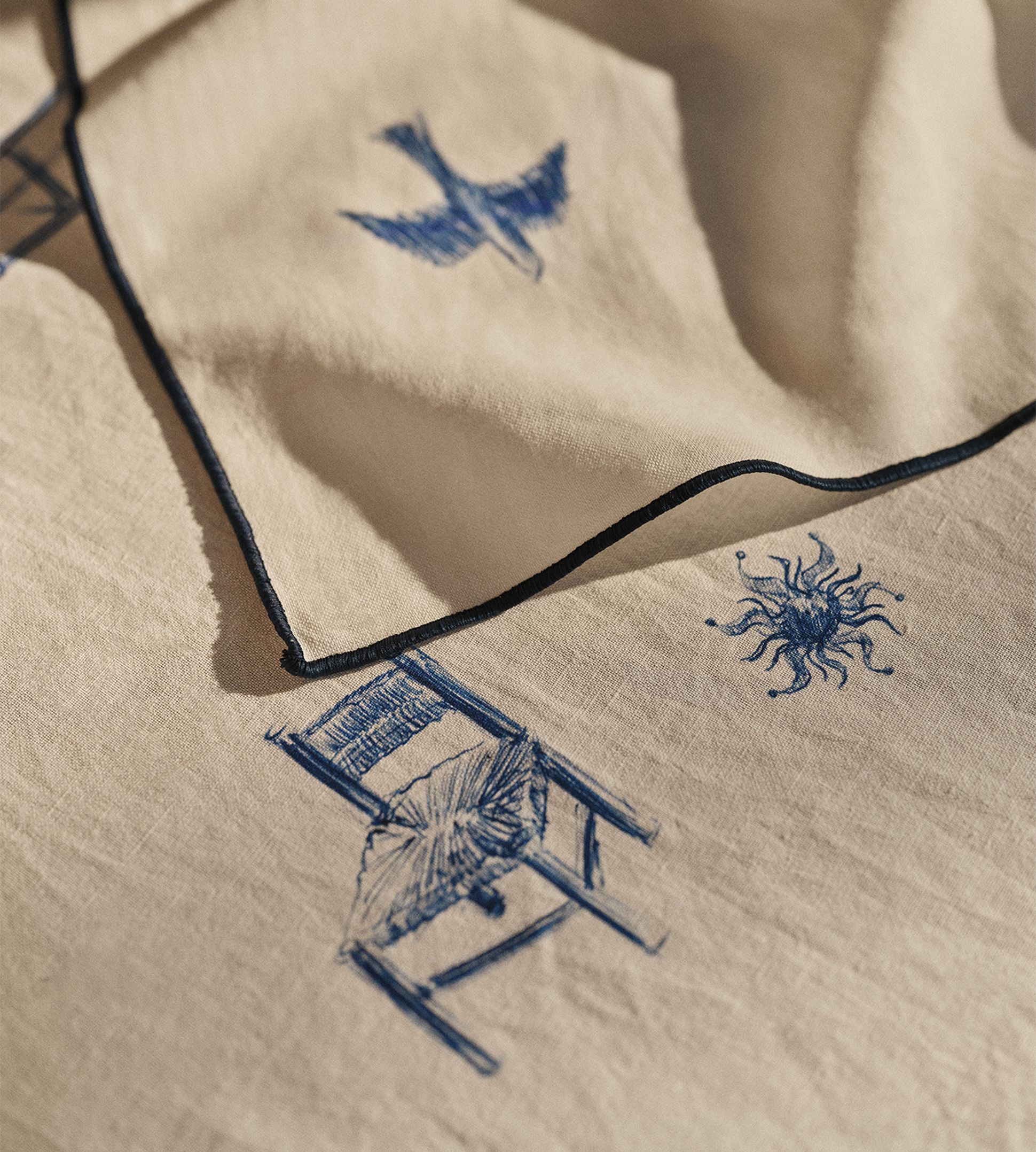 Gros plan sur un tissu beige orné de motifs brodés bleus représentant un oiseau, une forme ressemblant à un soleil et une chaise, le tout bordé d'une passementerie foncée.