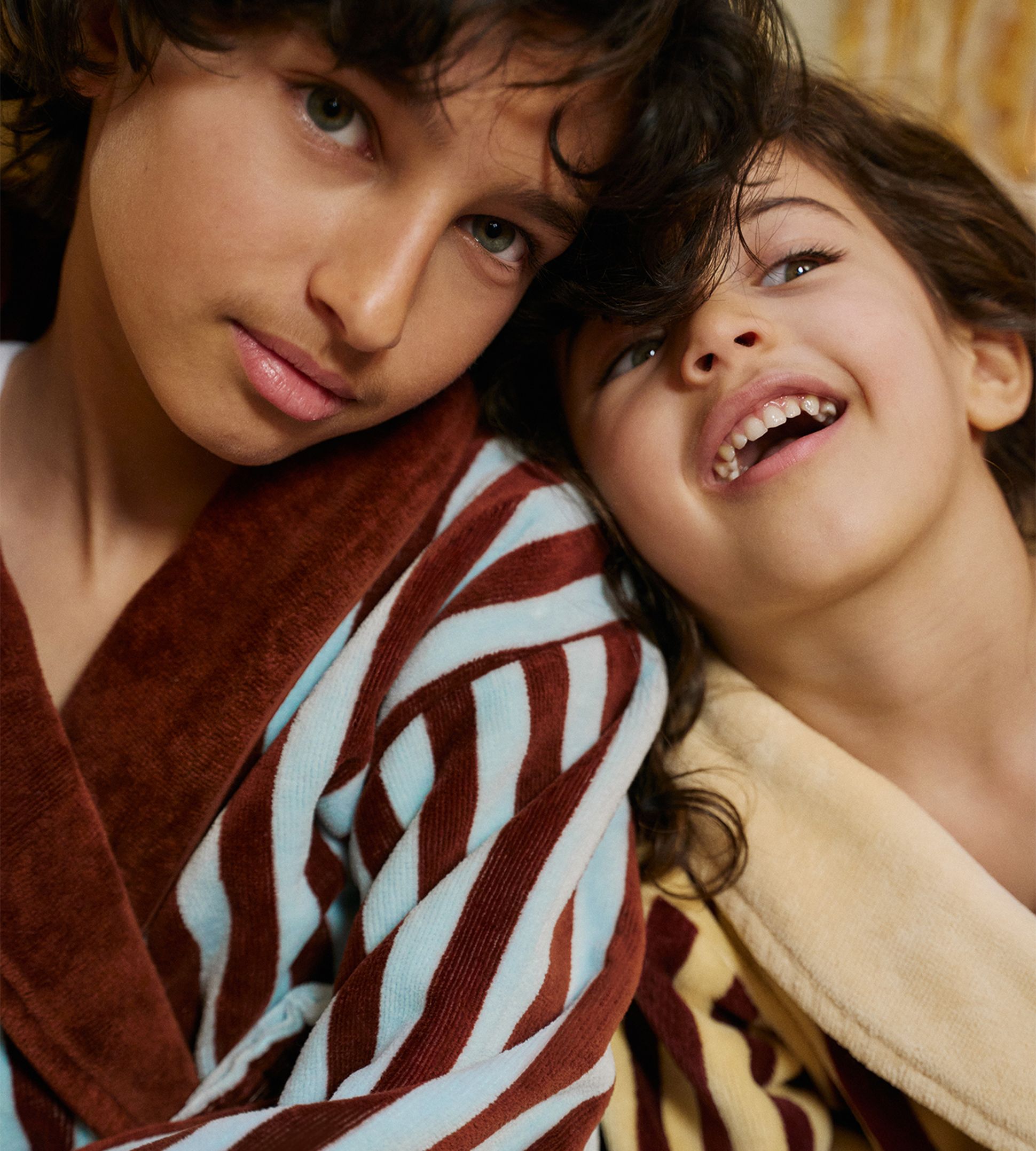 Deux enfants vêtus de peignoirs rayés bien douillets, l'un bleu et marron, l'autre jaune et marron, partagent un moment de joie et de complicité.