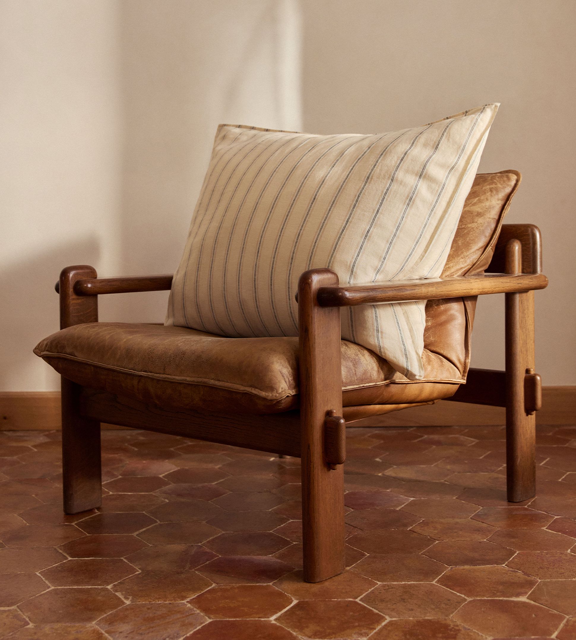 Un fauteuil en cuir à structure en bois, orné d'un coussin rayé, posé sur un sol en carrelage hexagonal dans une pièce baignée d'une lumière tamisée.