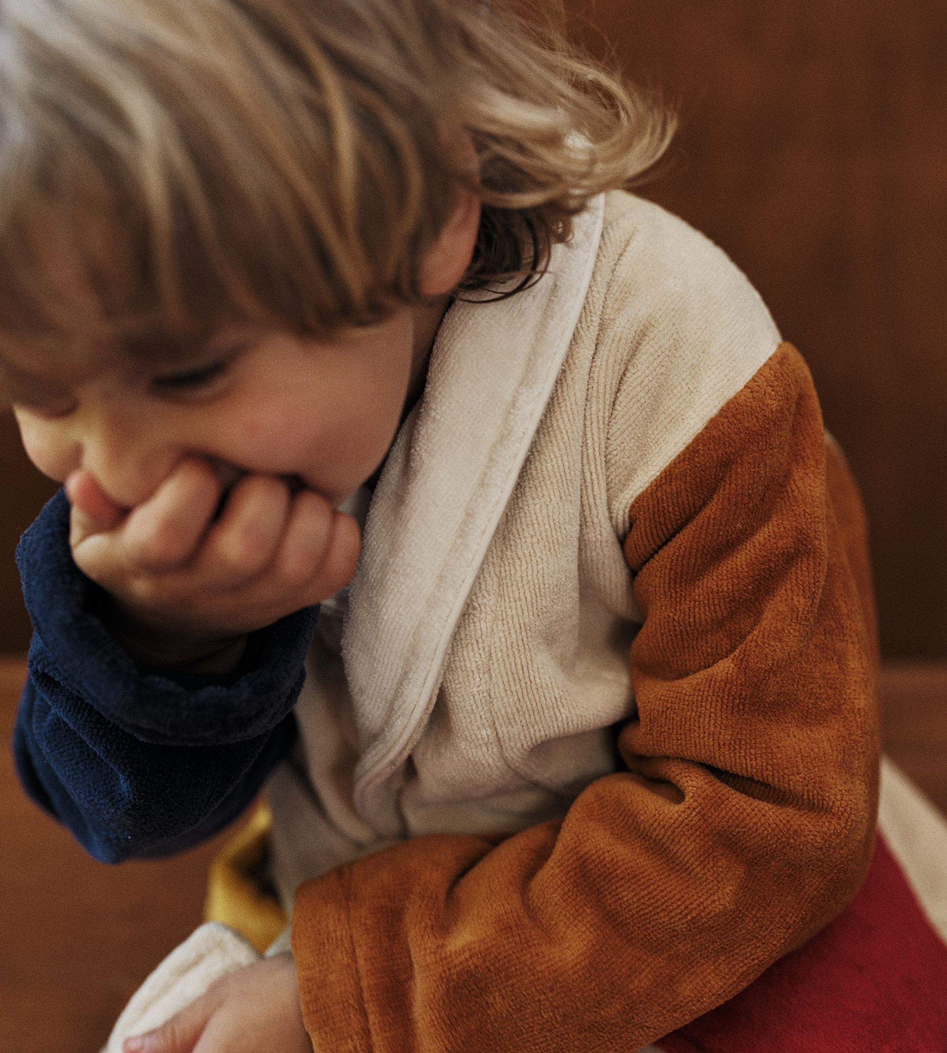 Un enfant aux cheveux blonds, vêtu d'une robe colorée, se couvre la bouche d'une main et semble joyeux dans un cadre chaleureux.