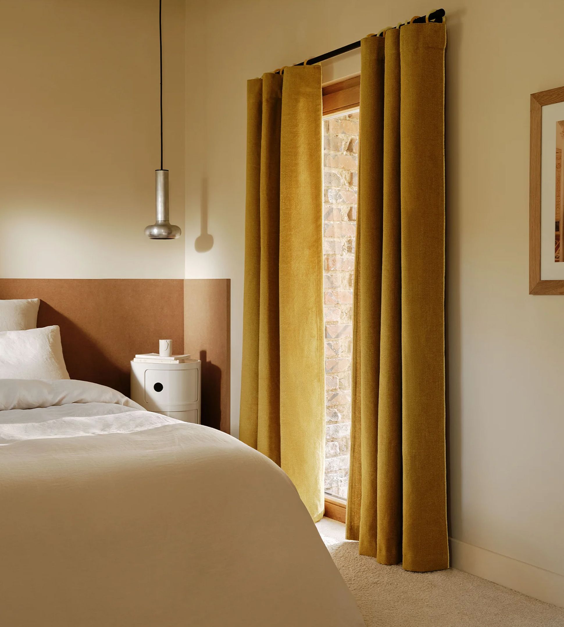 Chambre à coucher chaleureuse avec un lit garni de draps blancs, une petite table de chevet ronde, une suspension argentée et des rideaux jaune moutarde devant la fenêtre.