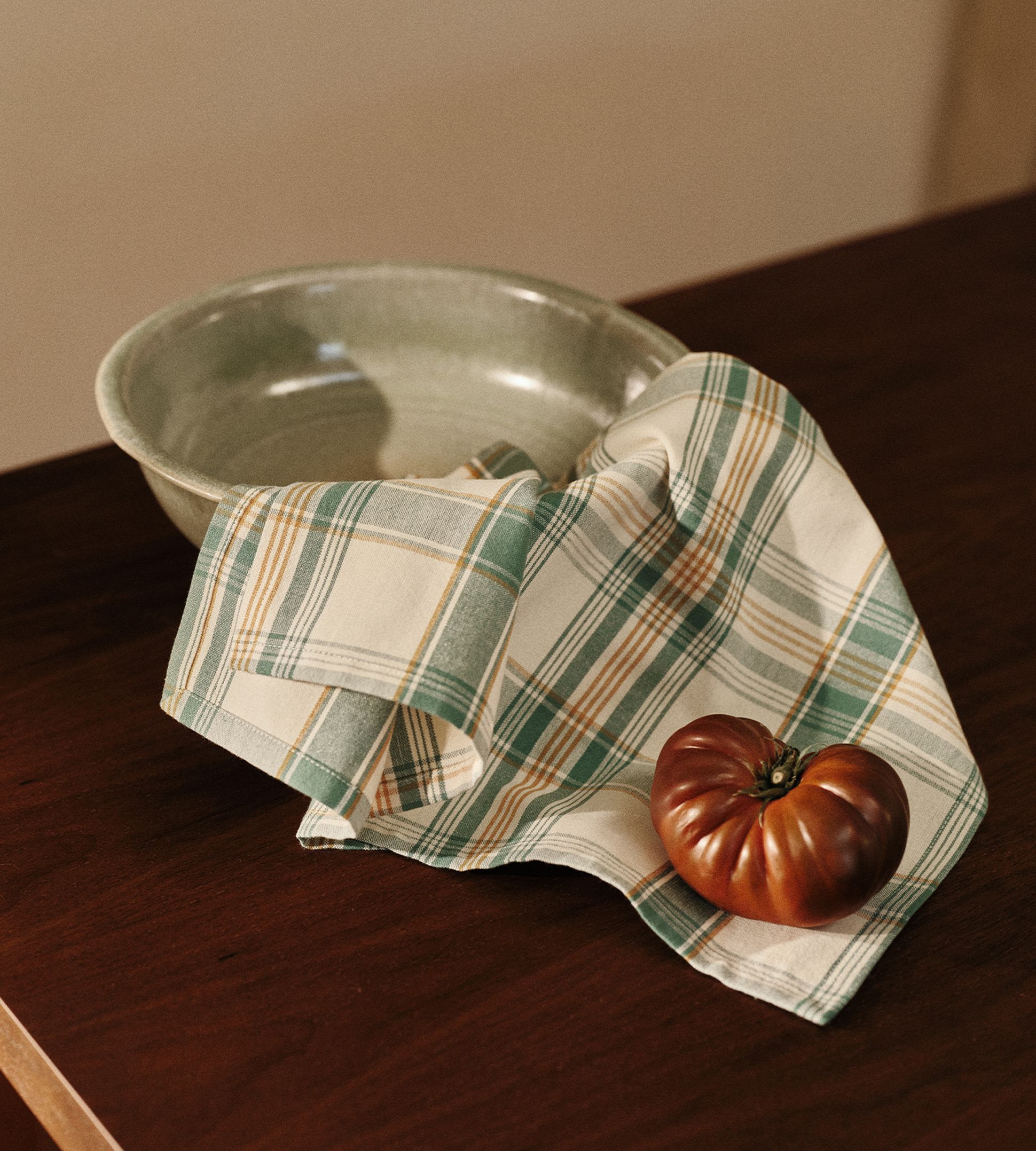 Un bol en céramique verte et un torchon rayé sur lequel repose une tomate, posés sur une table en bois.