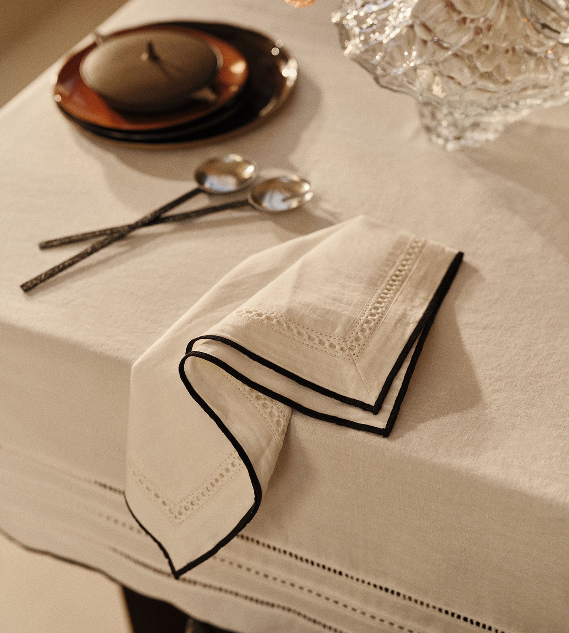 Une table élégamment dressée, avec une serviette blanche pliée aux bords noirs, deux cuillères, des assiettes empilées et un plat décoratif en verre, le tout baigné d'une lumière tamisée.