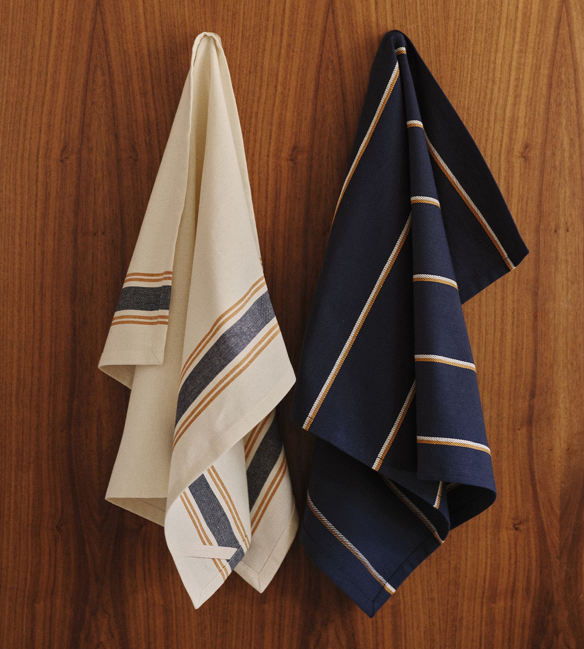 Deux torchons rayés, l'un beige et l'autre bleu marine, suspendus à un mur en bois.