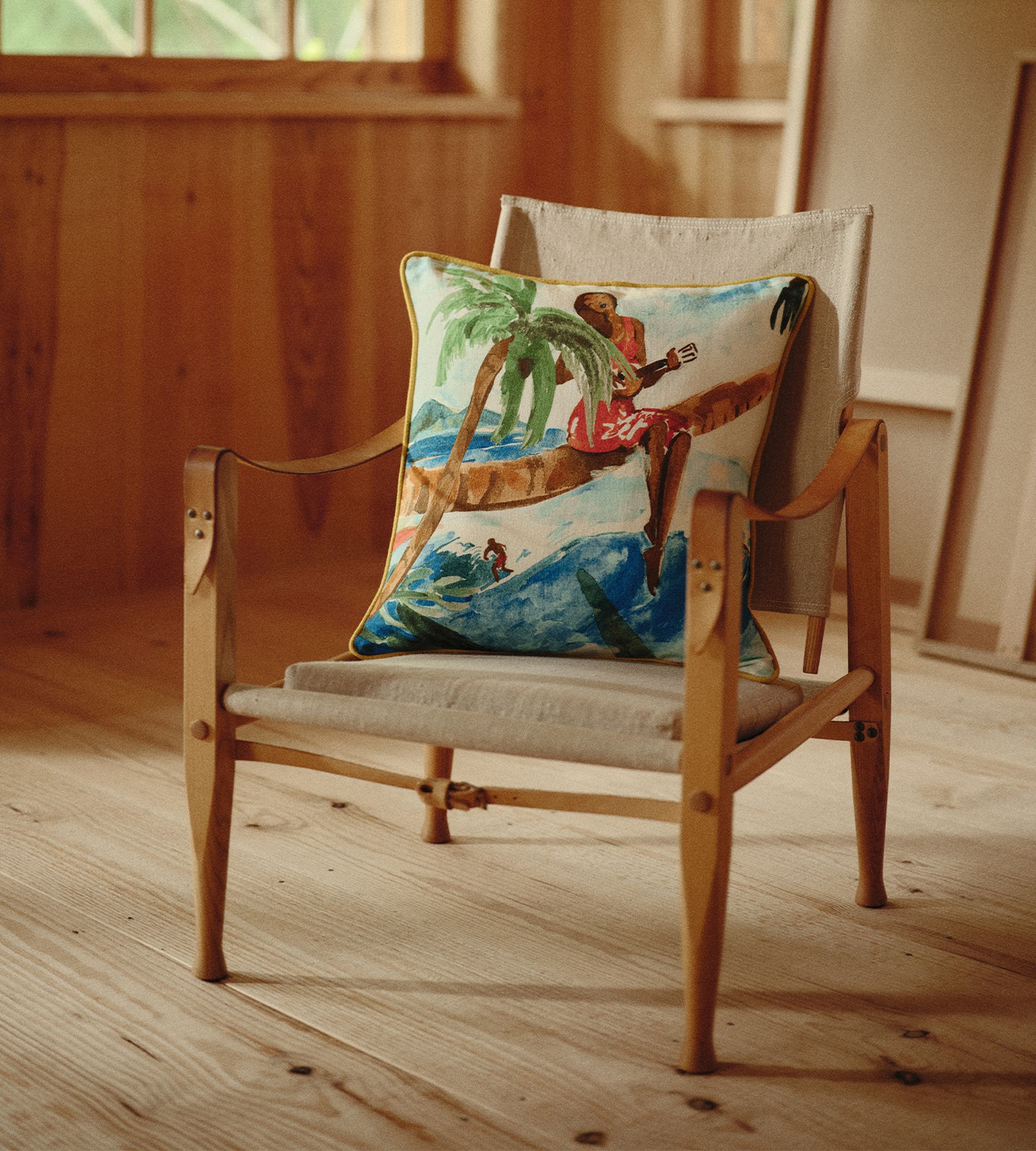 Chaise en bois avec un coussin coloré représentant un paysage tropical, dans une pièce baignée de soleil aux murs et au sol en bois.