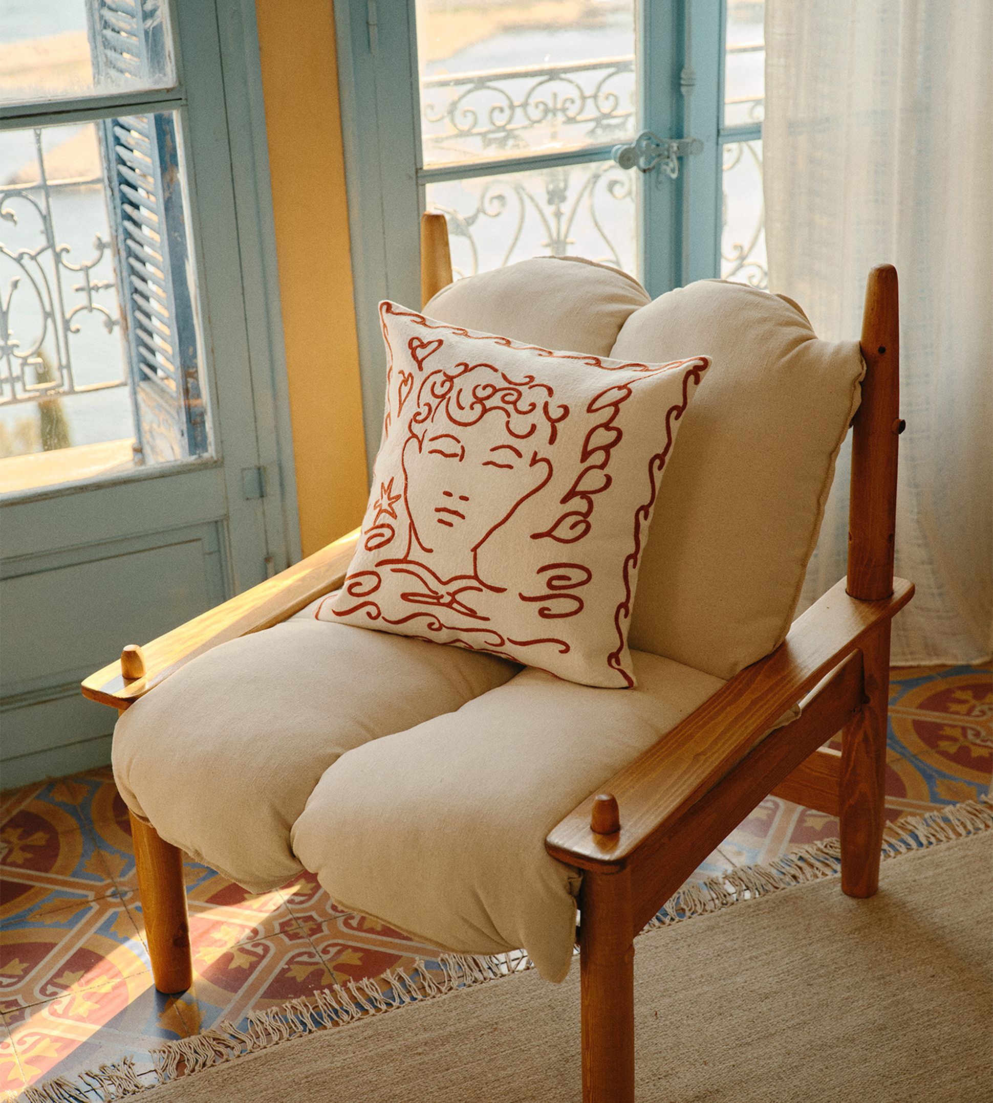 Un fauteuil confortable avec des coussins beiges et un coussin décoratif orné d'un visage dessiné au trait rouge, placé près d'une fenêtre aux encadrements bleu clair et d'un sol en carrelage aux motifs complexes.
