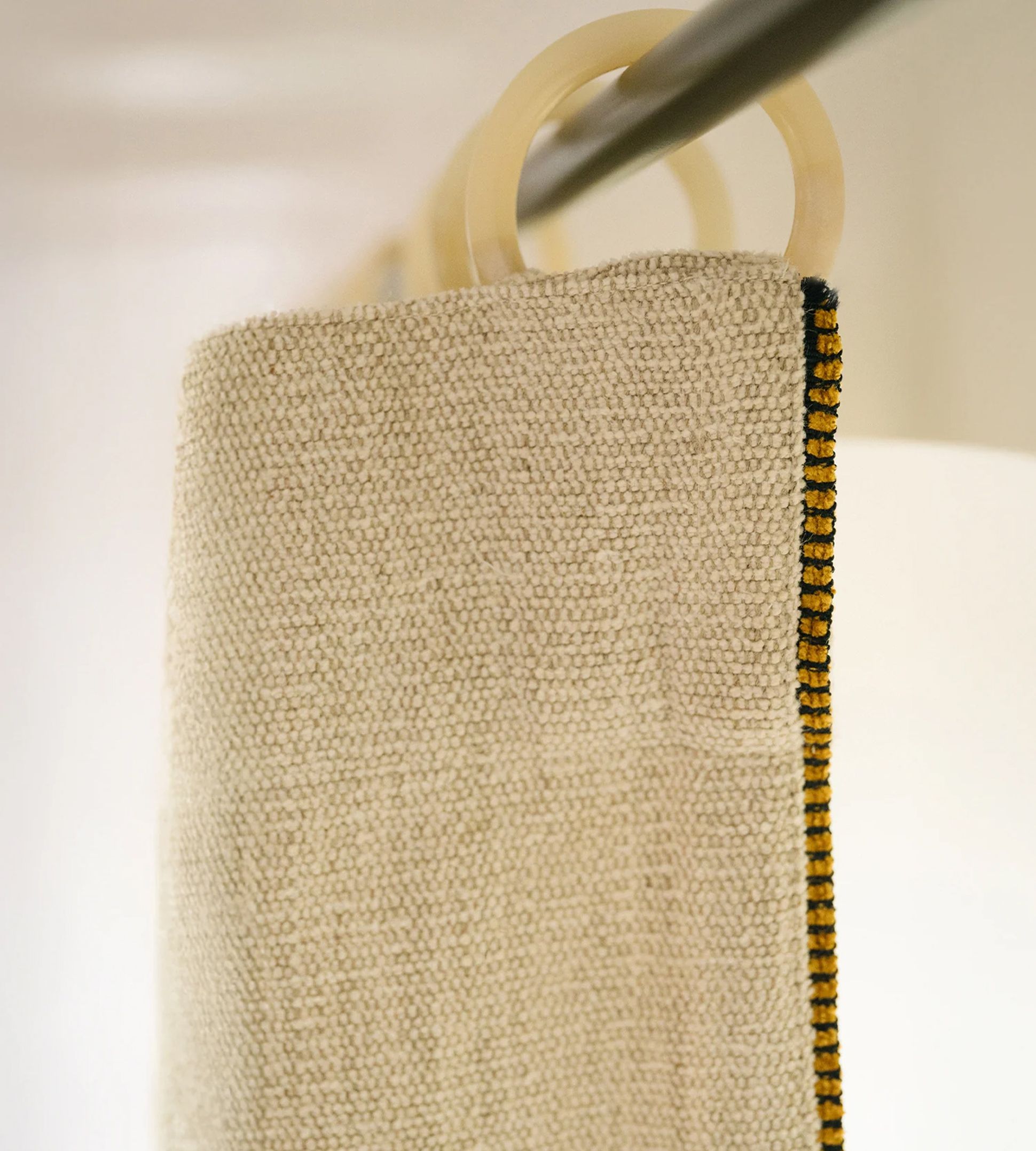 Gros plan sur un rideau beige texturé, orné d'une bordure noire et jaune, suspendu à des anneaux blancs fixés à une tringle métallique.