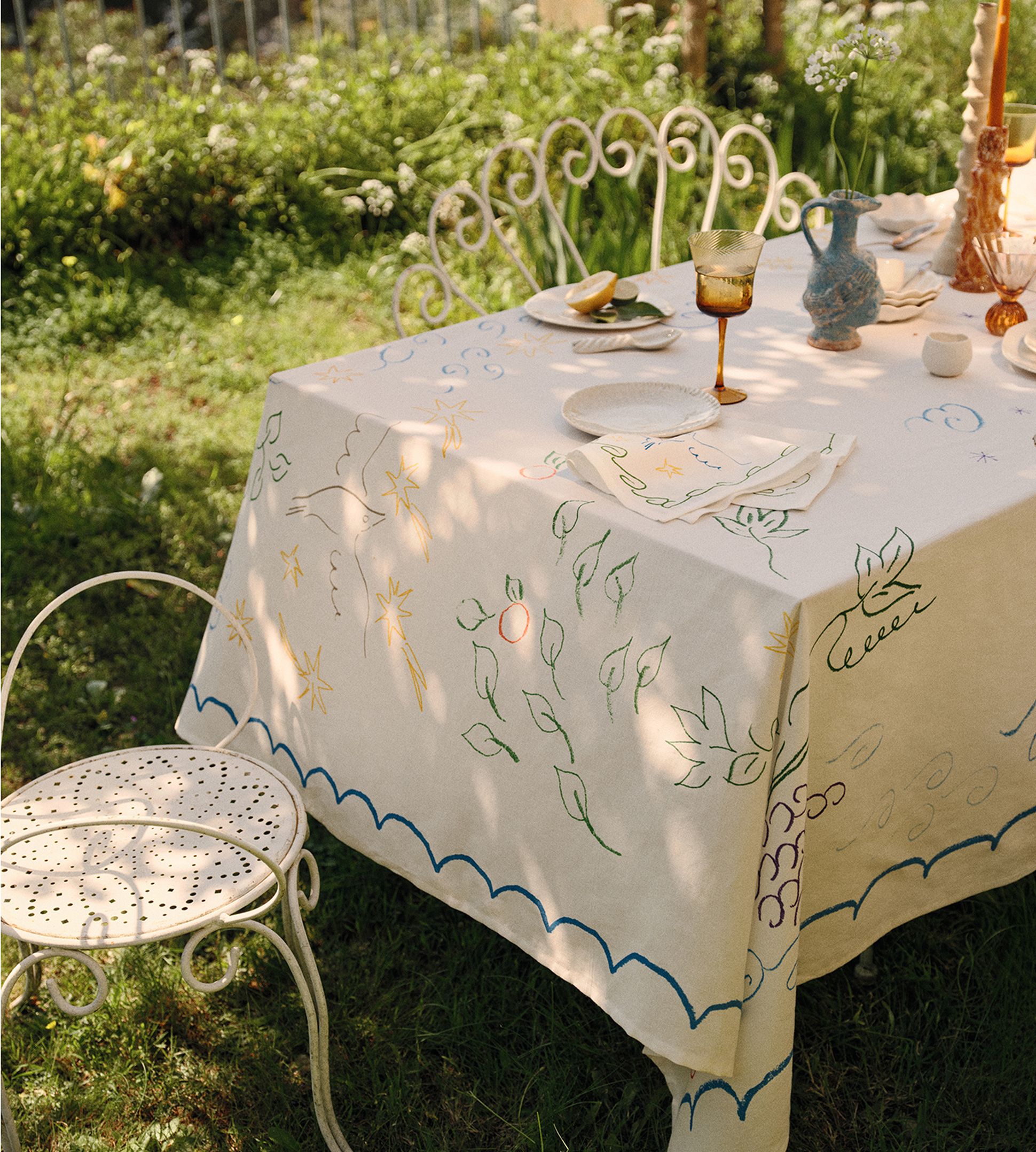Une table dressée en plein air, recouverte d'une nappe blanche ornée de motifs colorés représentant des feuilles et des étoiles, entourée d'élégantes chaises blanches dans un jardin.