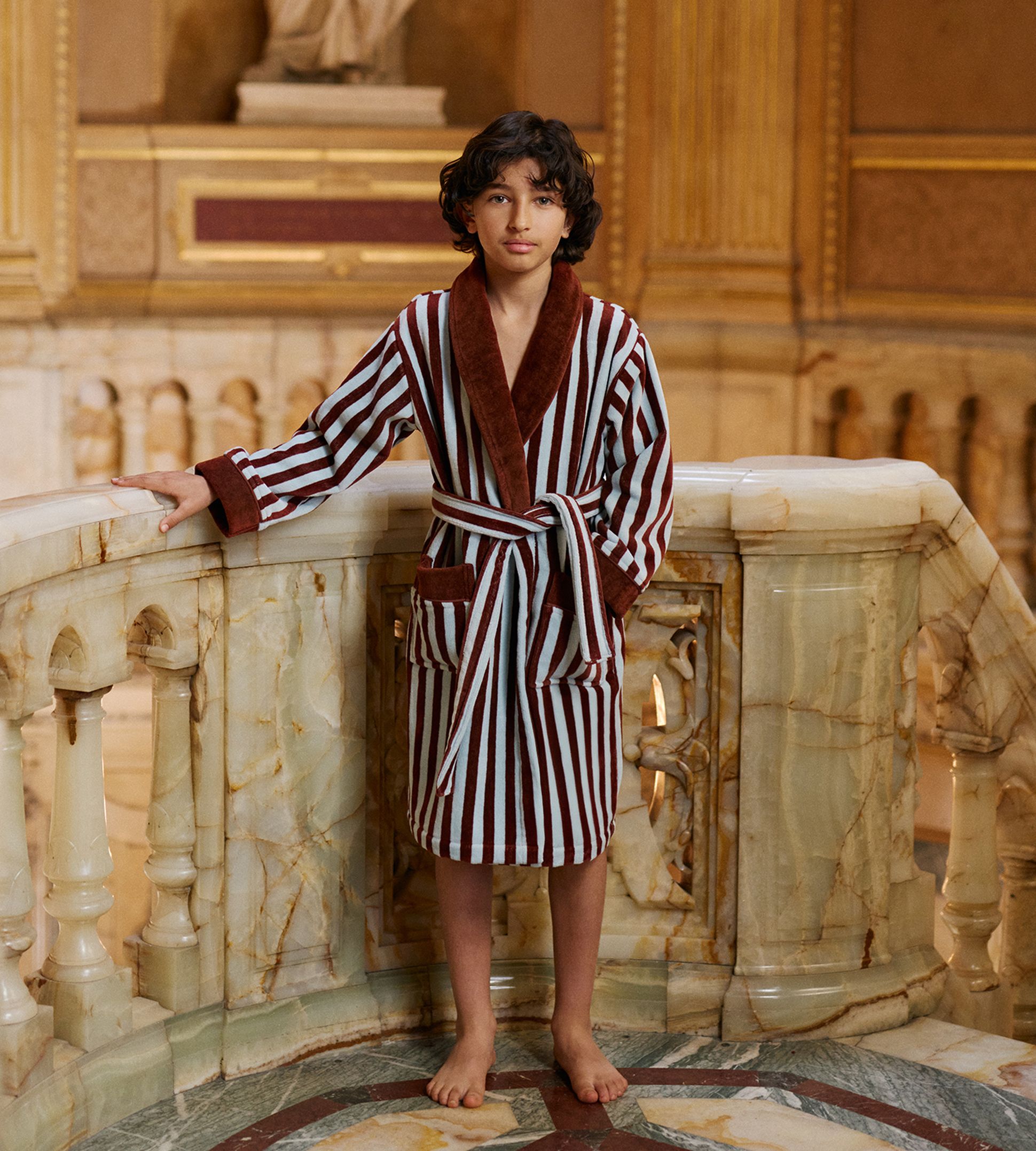 Un enfant vêtu d'une robe rayée se tient pieds nus sur un sol en marbre richement décoré, avec une statue classique en arrière-plan.