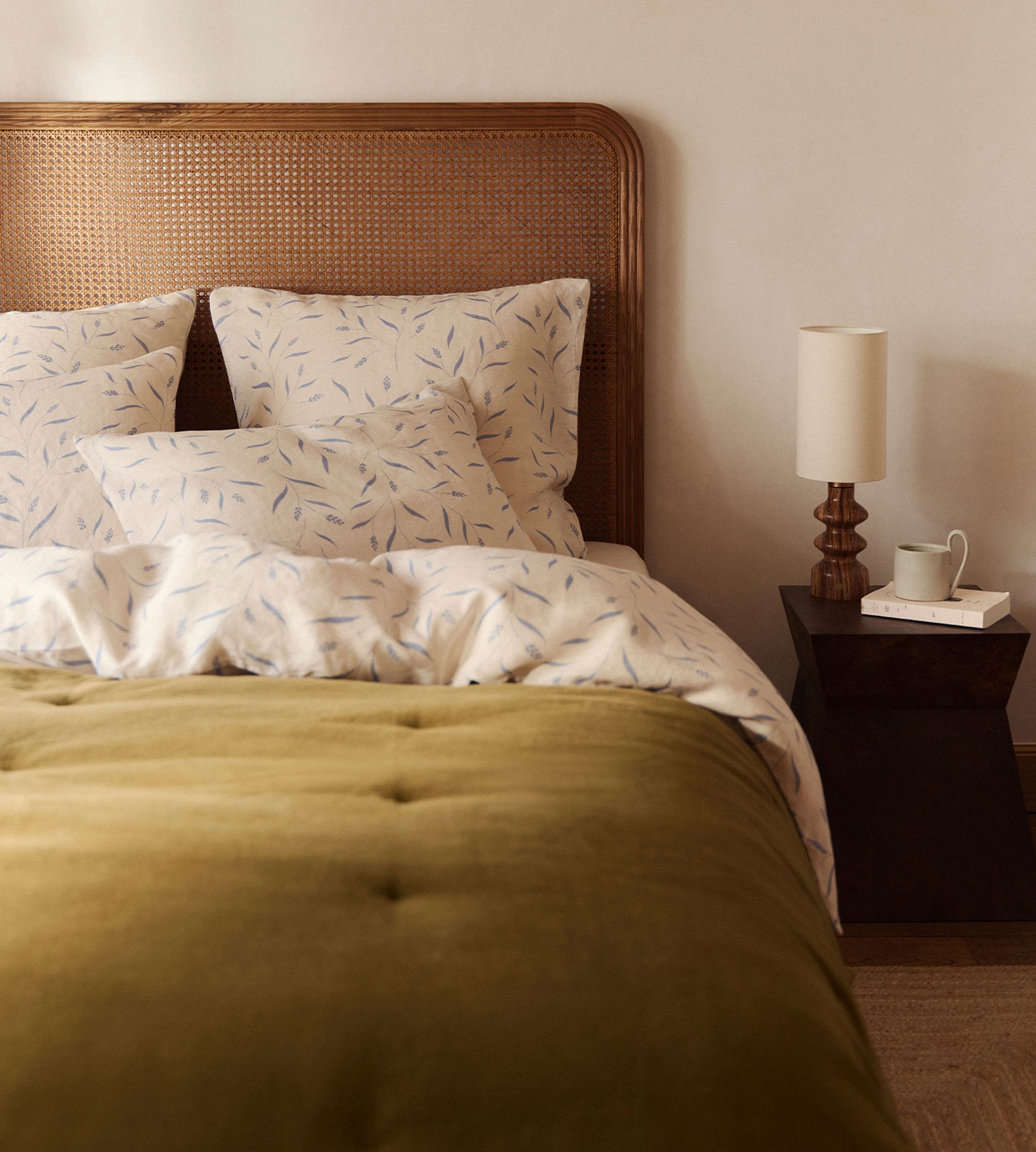 Une chambre douillette avec une tête de lit en bois, des coussins à motifs, une couverture vert olive et une table de chevet sur laquelle sont posés une lampe, des livres et une tasse.