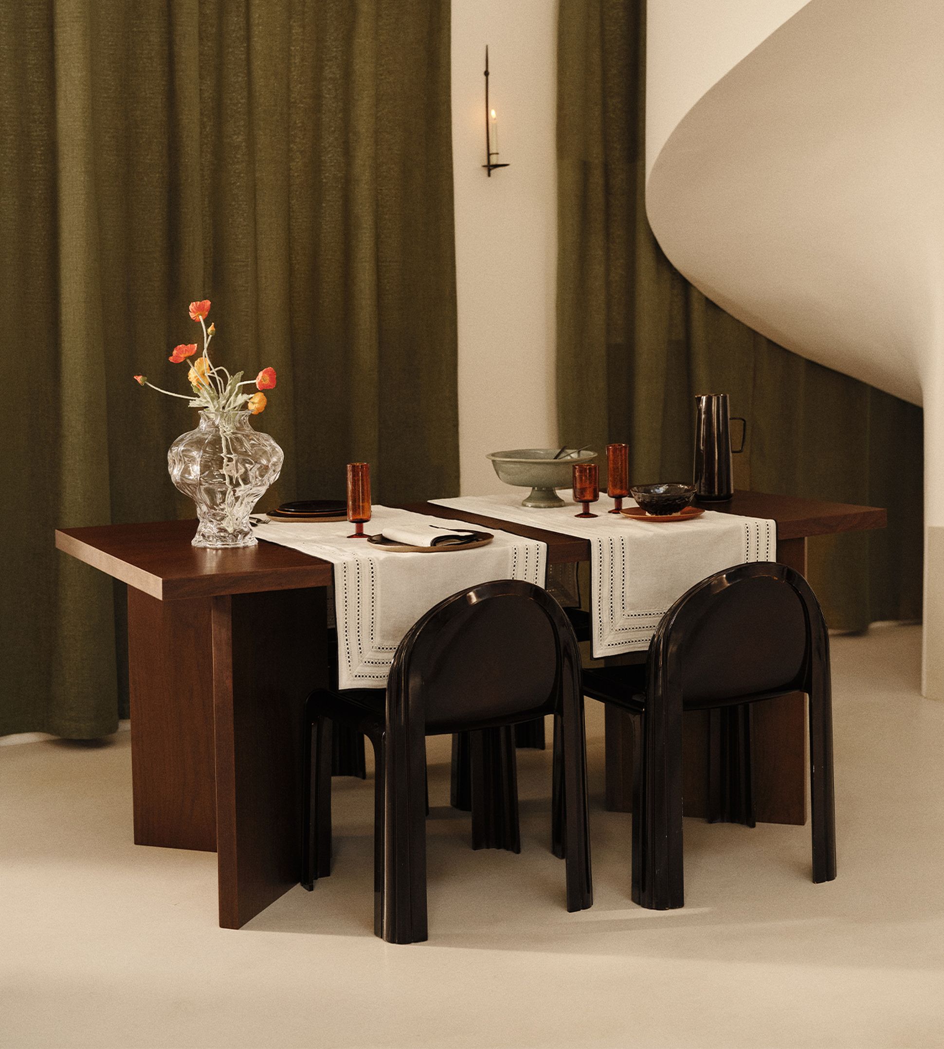 Une salle à manger élégante, avec une table en bois foncé, des chaises noires, un vase transparent rempli de fleurs et des verres ambrés posés sur des chemins de table blancs, le tout sur fond de rideaux vert olive.
