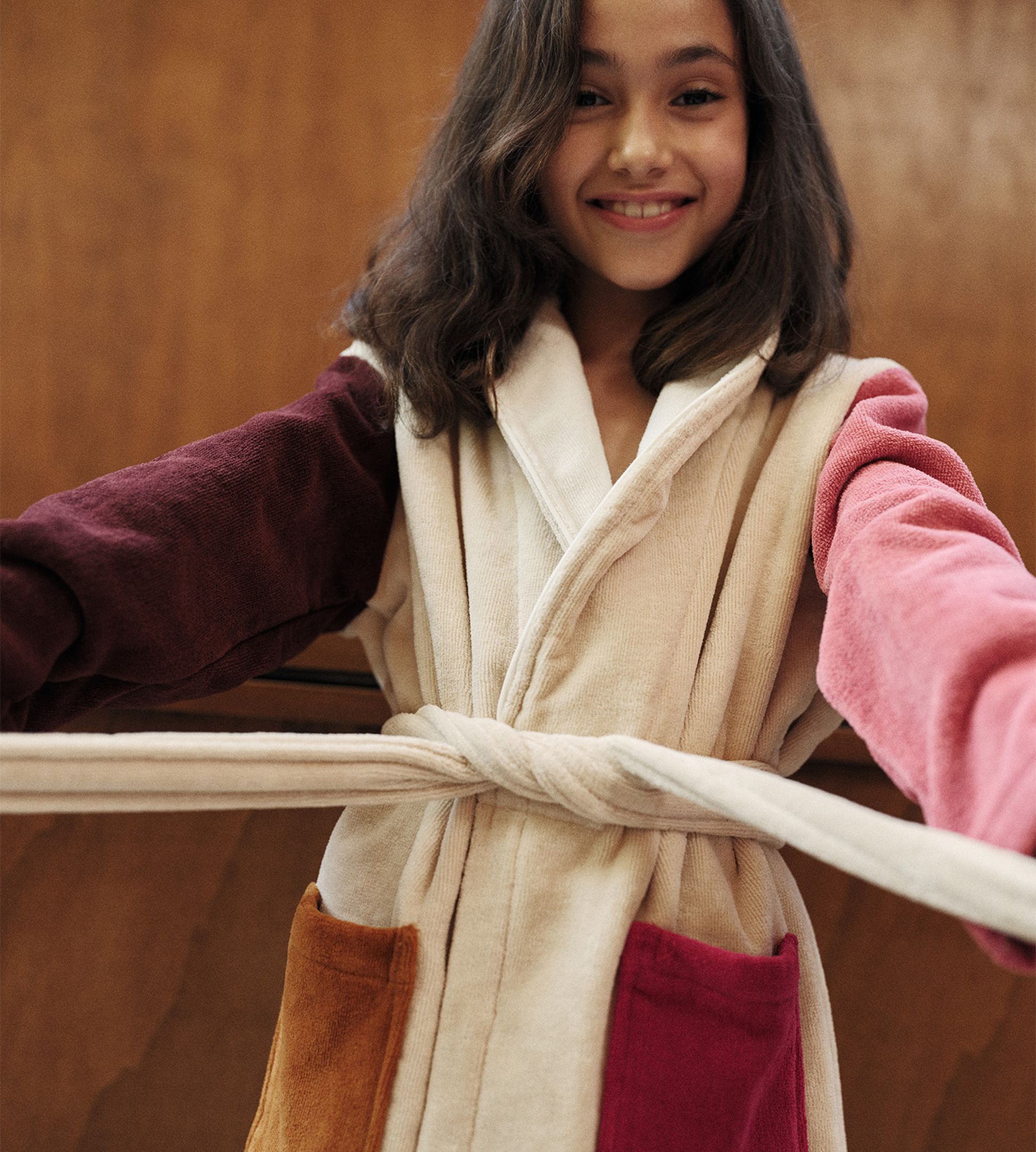 Une jeune fille souriante, vêtue d'une robe beige aux poches et aux manches colorées, debout devant un fond en bois.