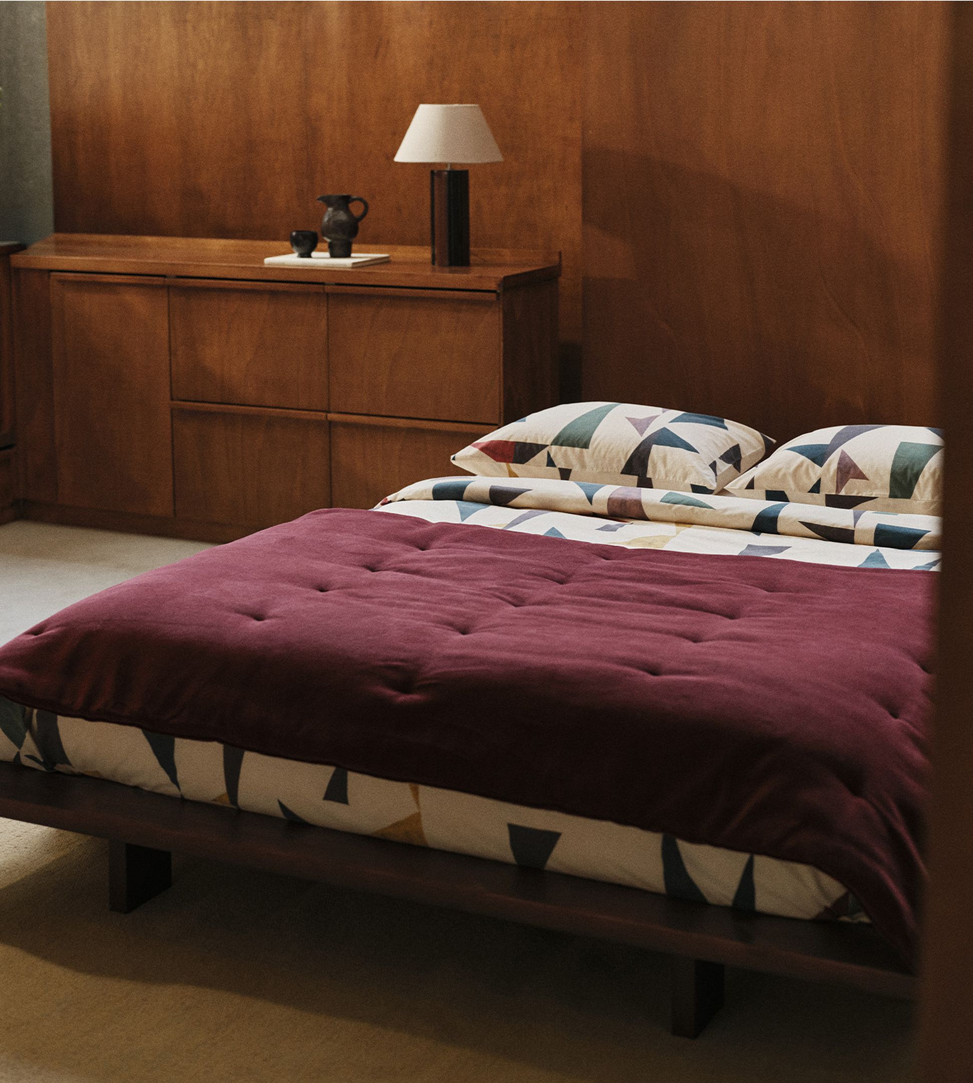 Une chambre en bois avec une couverture bordeaux sur le lit, des coussins aux motifs colorés, des commodes en bois, une lampe et de grandes fenêtres donnant sur la verdure.