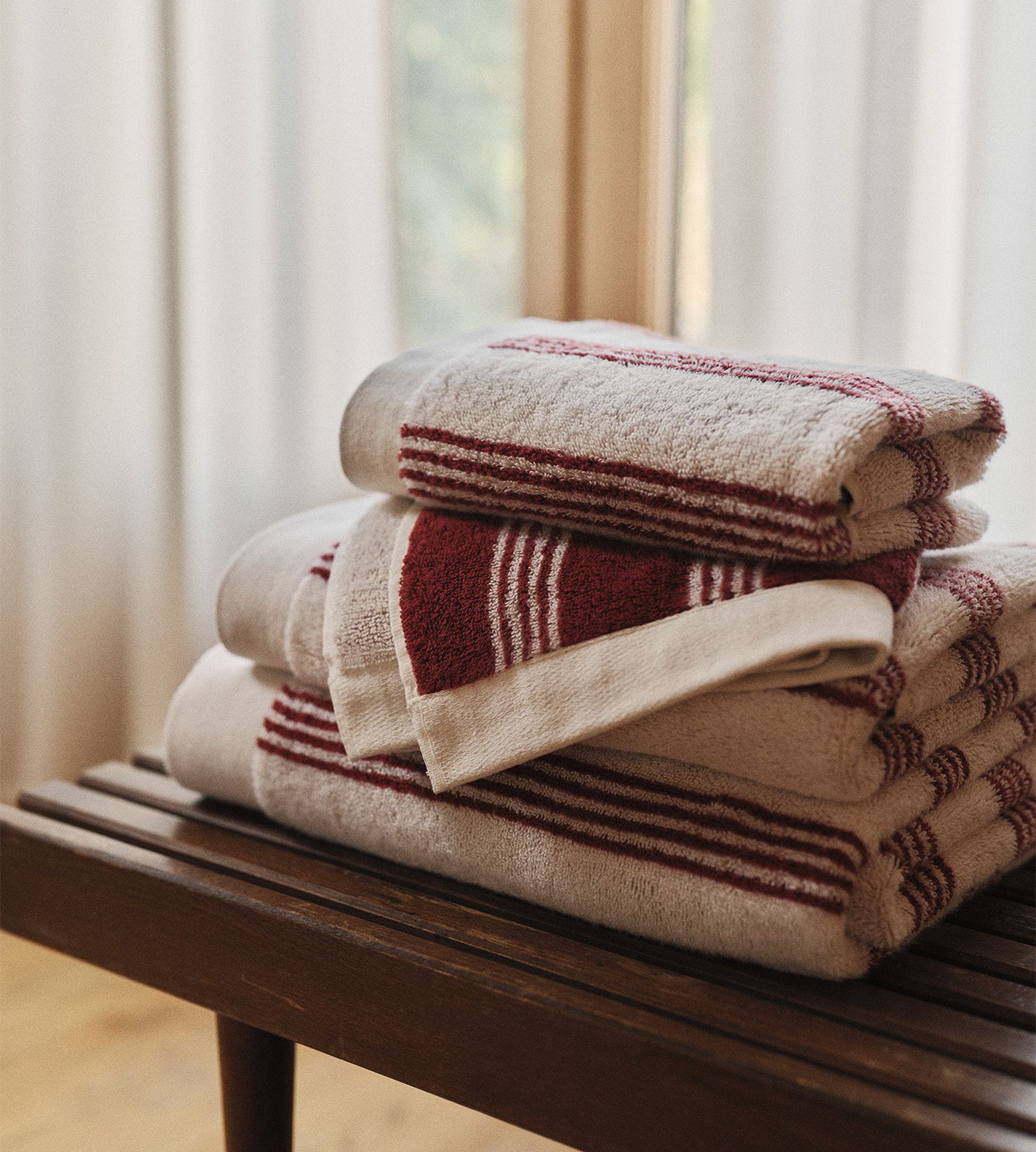 Une pile de serviettes rayées de beige et de rouge posée sur un banc en bois, avec une douce lumière naturelle qui filtre à travers une fenêtre à l'arrière-plan.