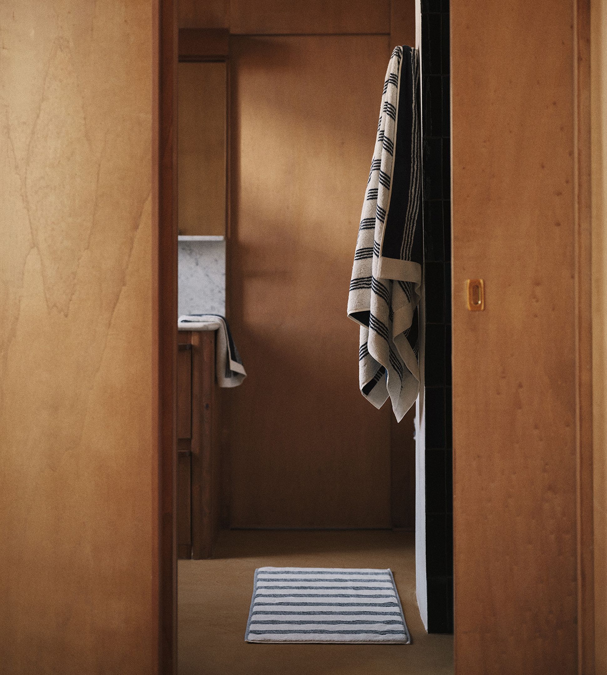 Une salle de bains chaleureuse, avec ses murs en bois, ses serviettes rayées suspendues et son tapis rayé au sol, qui créent une ambiance chaleureuse et minimaliste.