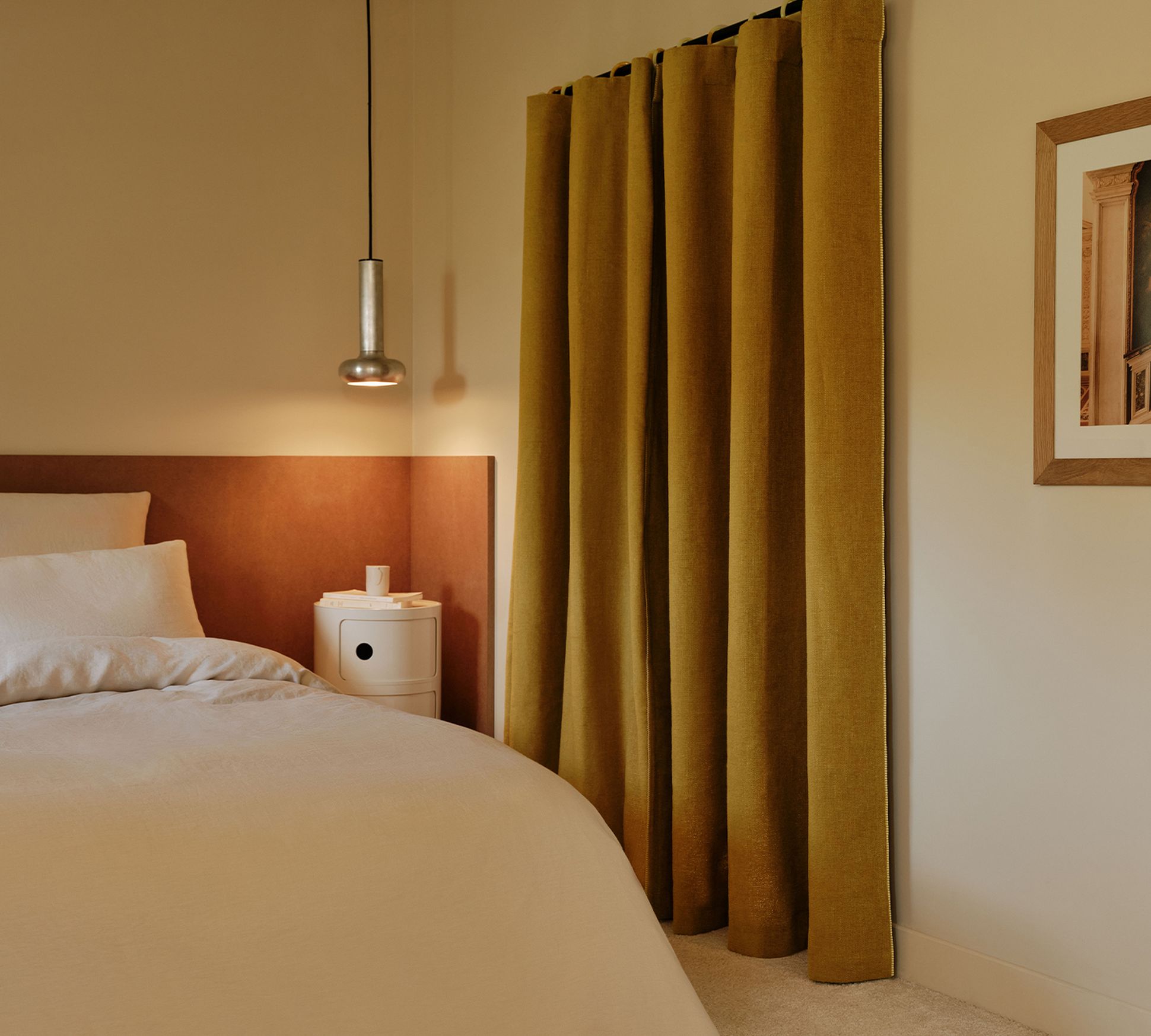 Cozy bedroom with a bed, white bedding, mustard curtains, a pendant light, and a small round bedside table with a framed picture on the wall.