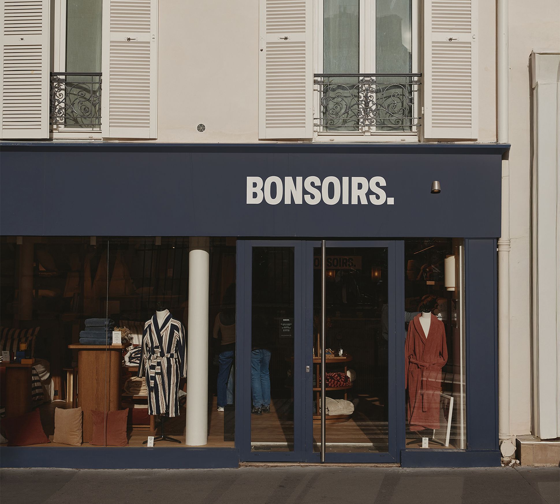 Vue de rue d'un magasin de vêtements à la façade bleu marine portant l'enseigne « BONSOIRS », avec des vêtements et des accessoires exposés en vitrine.