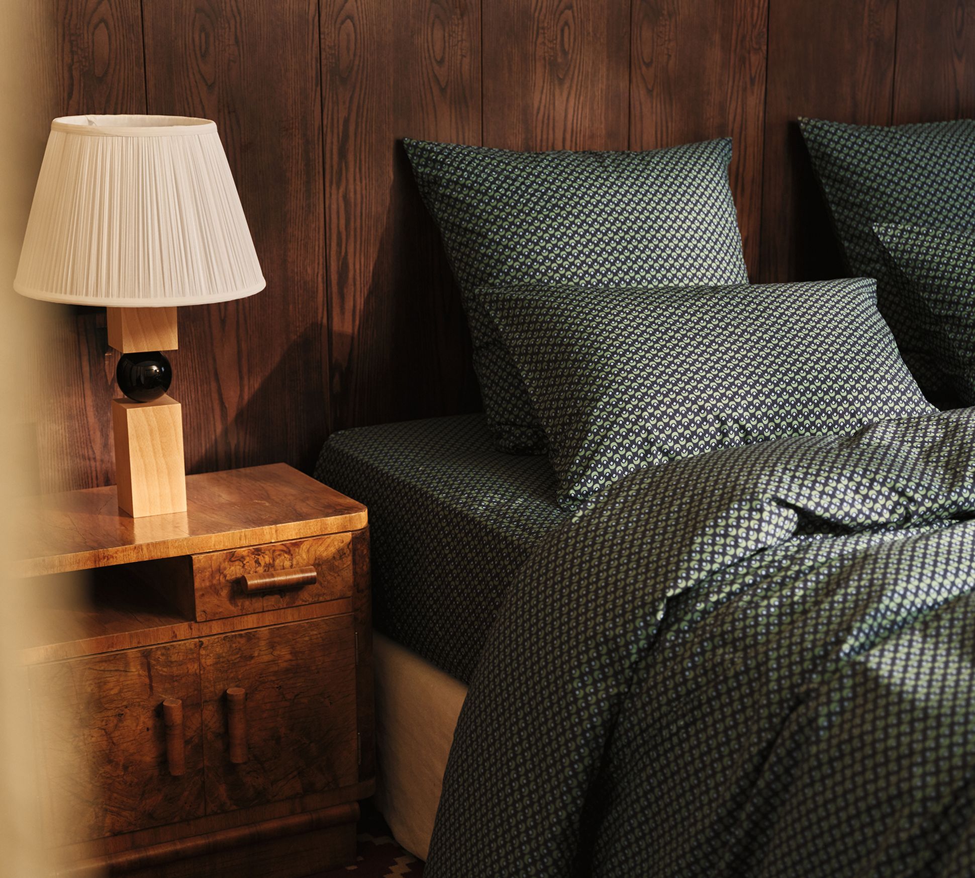 A cozy bedroom with a wooden nightstand, a table lamp, and a bed with green patterned bedding and pillows against a dark wood-paneled wall.