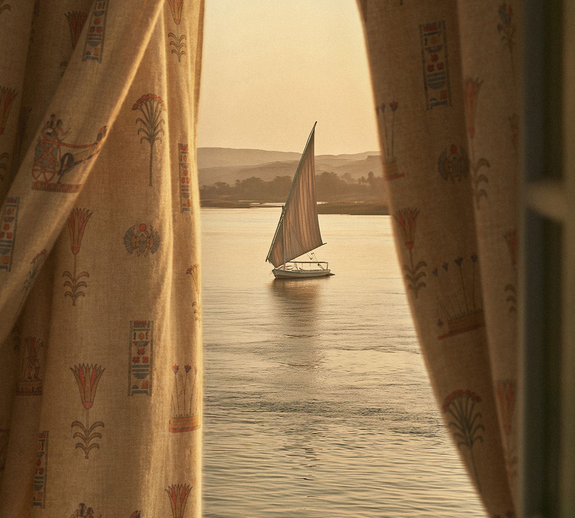A sailboat glides on a calm river at sunset, framed by curtains with traditional patterns, revealing a serene view.