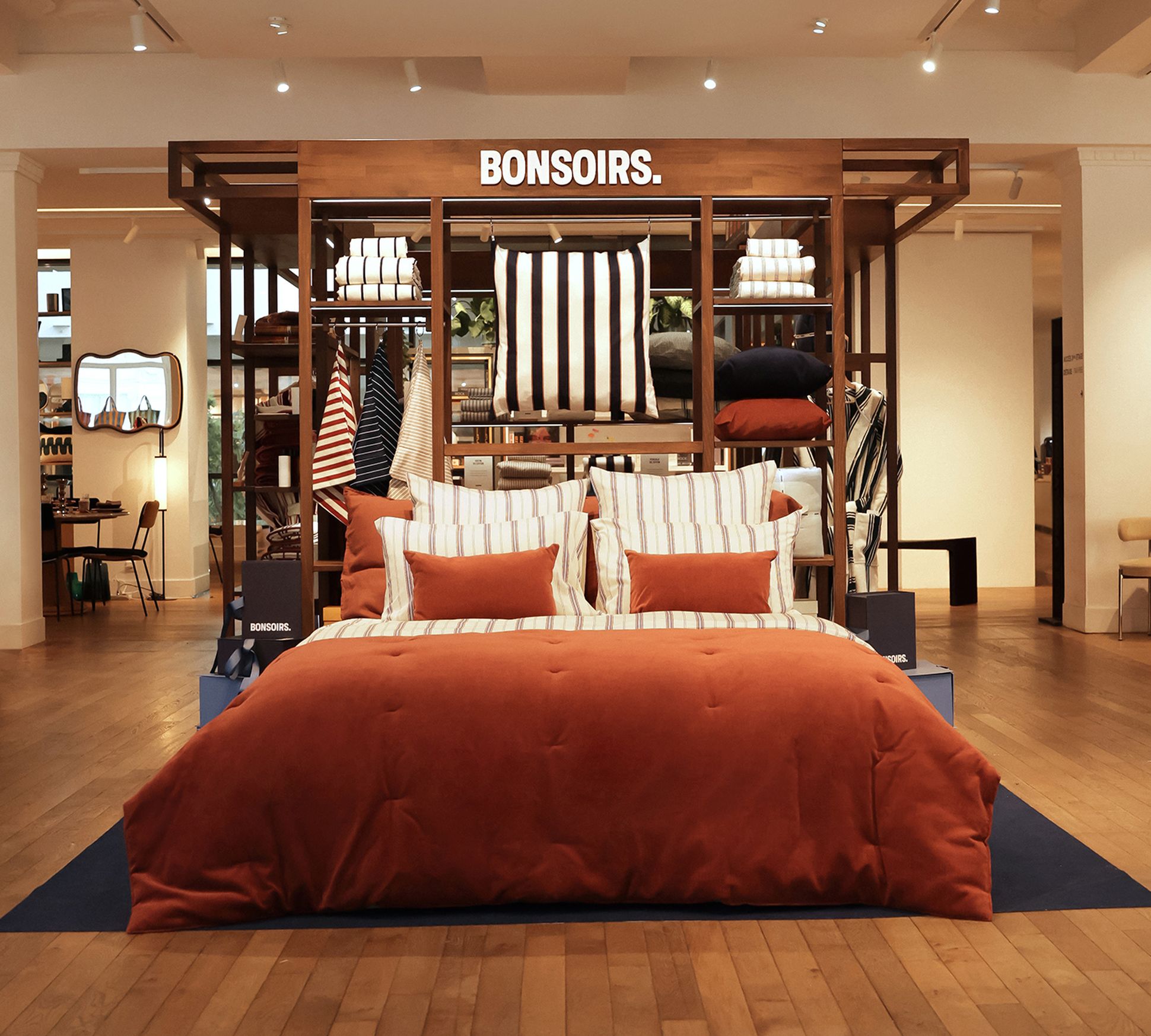 Dans un showroom chaleureux, on peut admirer un lit élégant, agrémenté d'une parure de lit couleur rouille et d'oreillers rayés, posé sur un parquet. Au-dessus, on aperçoit une enseigne « BONSOIRS. ».