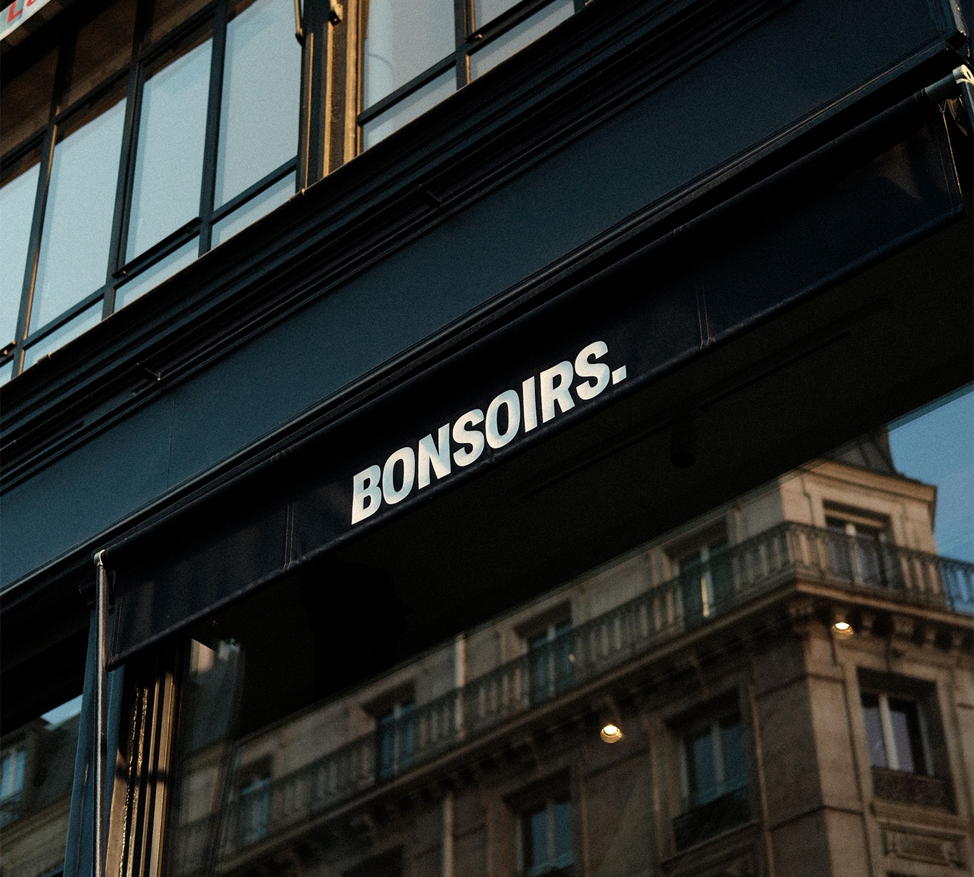 Une devanture avec un auvent bleu sur lequel est inscrit « BONSOIRS ». Les vitres réfléchissantes laissent entrevoir la façade d'un immeuble.