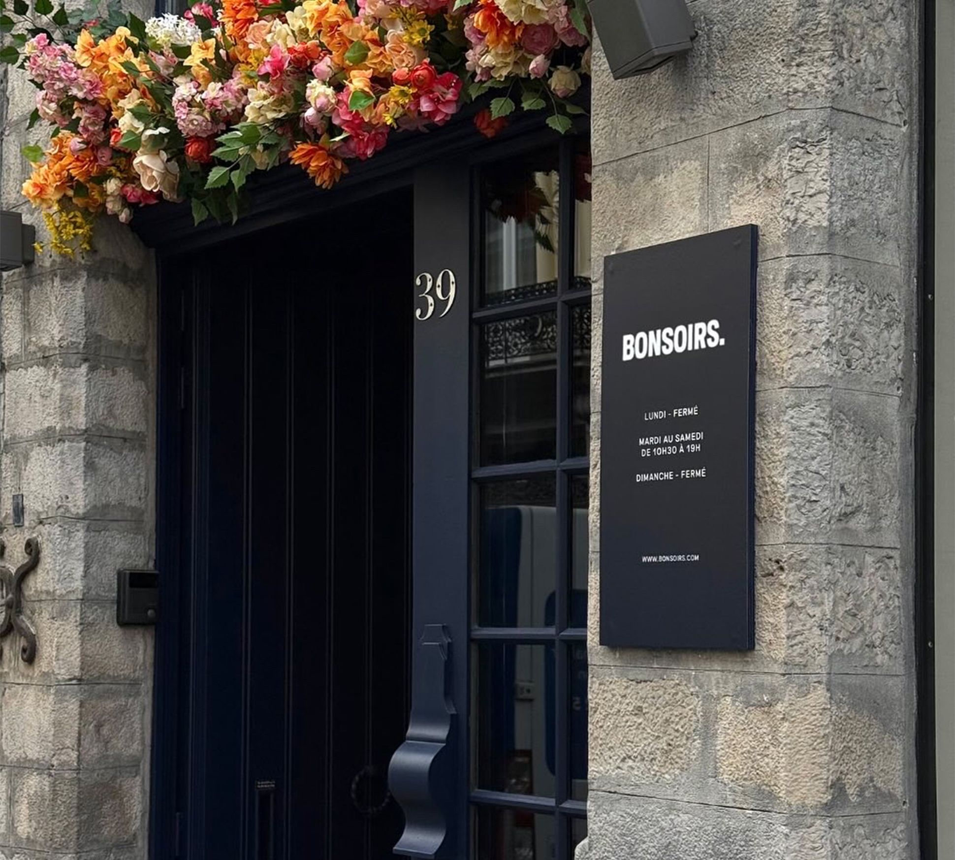 Porte bleu foncé surmontée d'une composition florale. Une enseigne indique « BONSOIRS » ainsi que les horaires d'ouverture. Façade en pierre sur laquelle on peut voir le numéro 39.