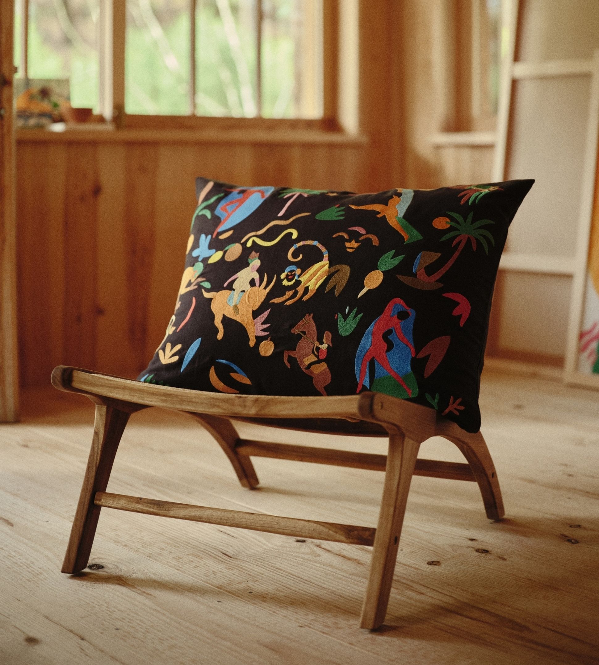 Un coussin coloré orné de motifs animaliers abstraits sur une chaise en bois, dans une pièce en bois baignée de soleil, dotée de grandes fenêtres et d'une lanterne en papier.