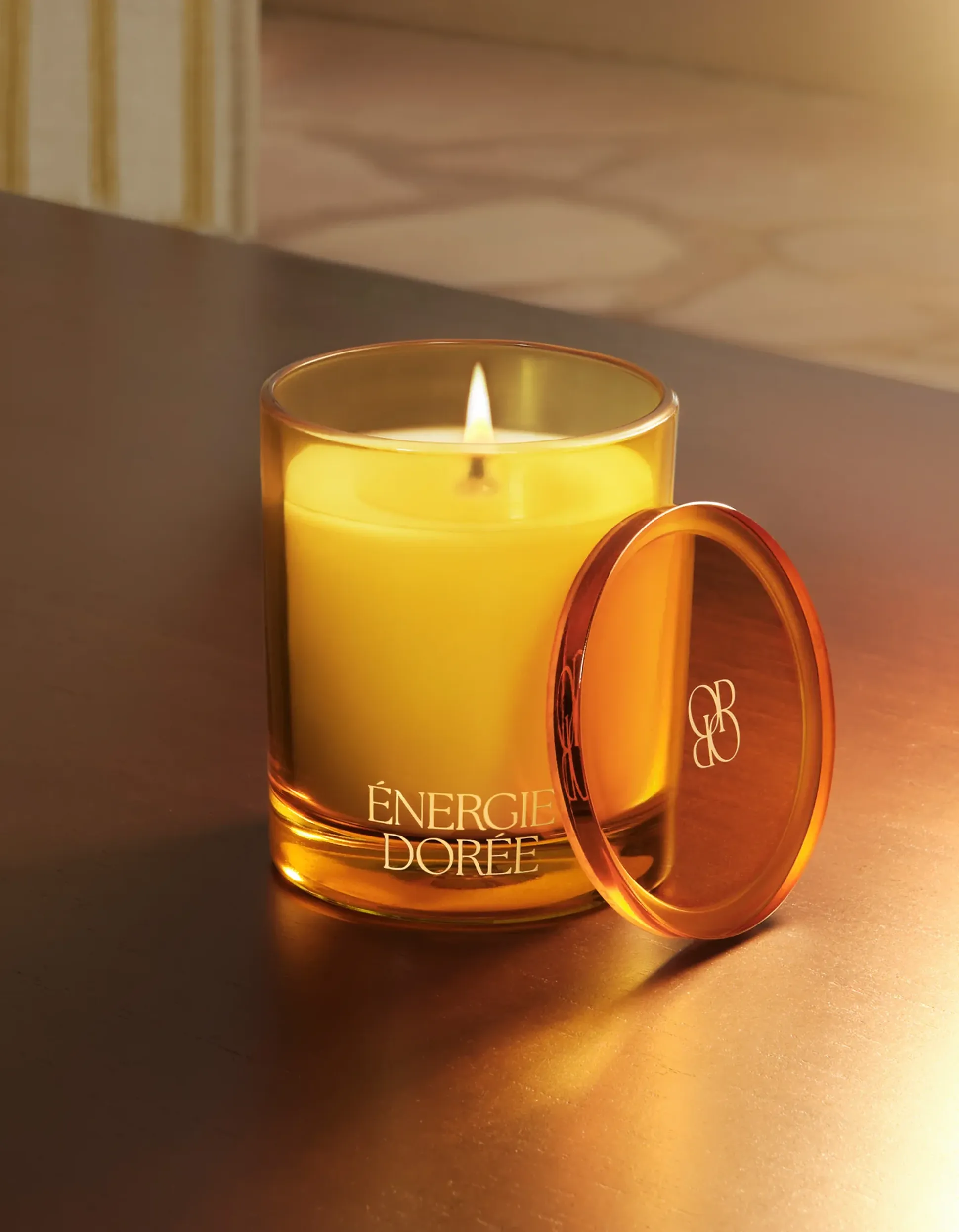 Une bougie jaune allumée dans un photophore en verre portant l'étiquette « ÉNERGIE DORÉE », avec son couvercle posé à côté, sur une surface en bois réfléchissante.