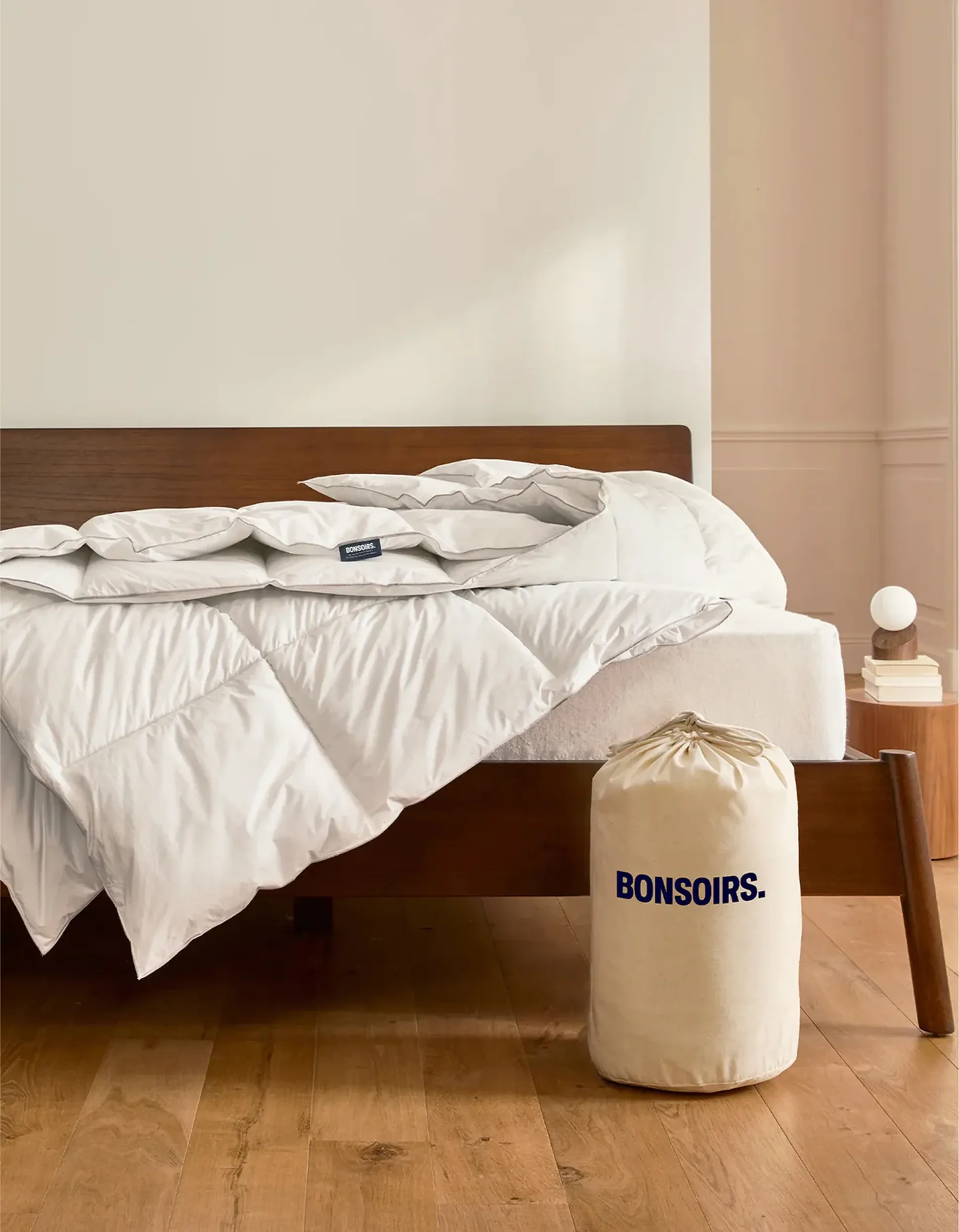 Un lit bien fait, recouvert d'une couette blanche moelleuse et d'un sac beige portant l'inscription « BONSOIRS », posé sur un parquet, à côté d'une table de chevet sur laquelle se trouvent une lampe et des livres.