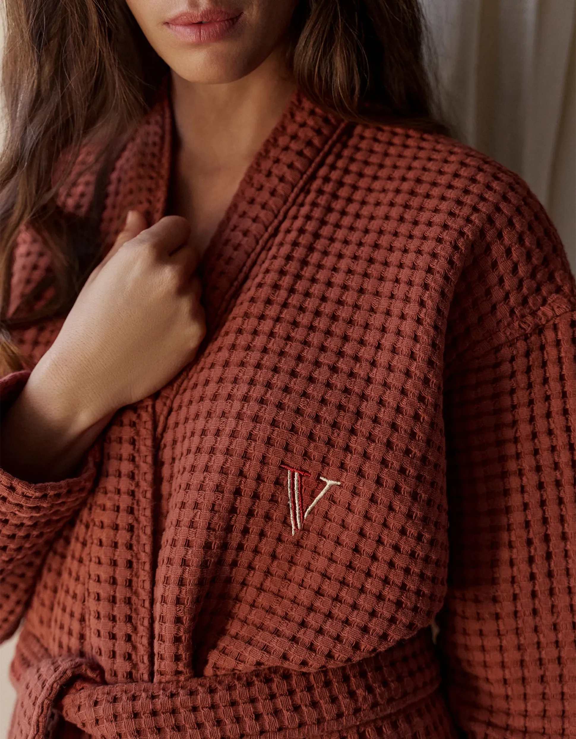 Femme portant un peignoir marron texturé à motif gaufré avec un logo « V » brodé, se tenant devant un rideau clair.