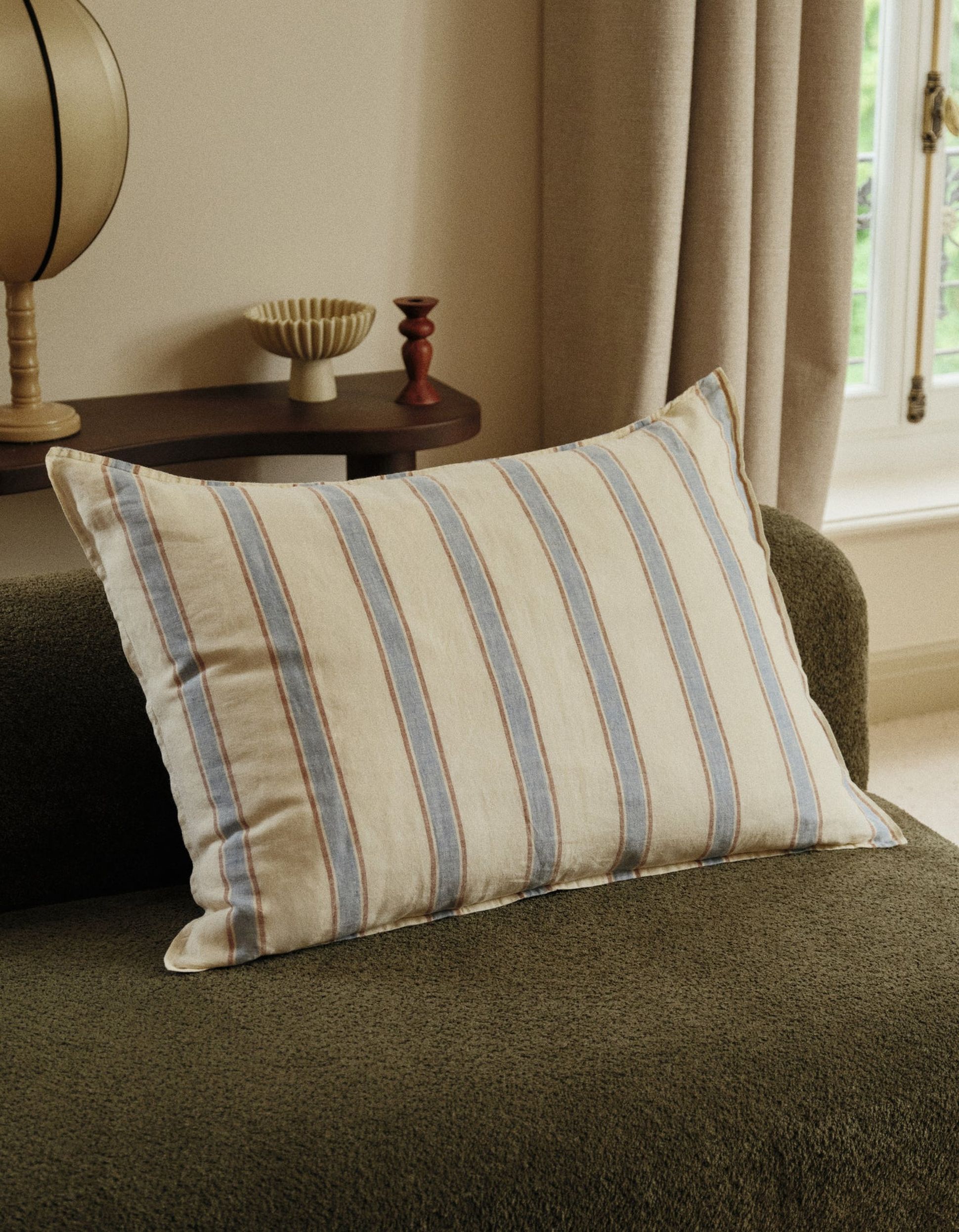 Un coussin rayé posé sur un canapé vert foncé, à côté d'une étagère ornée d'objets décoratifs, dans une pièce baignée d'une lumière tamisée.