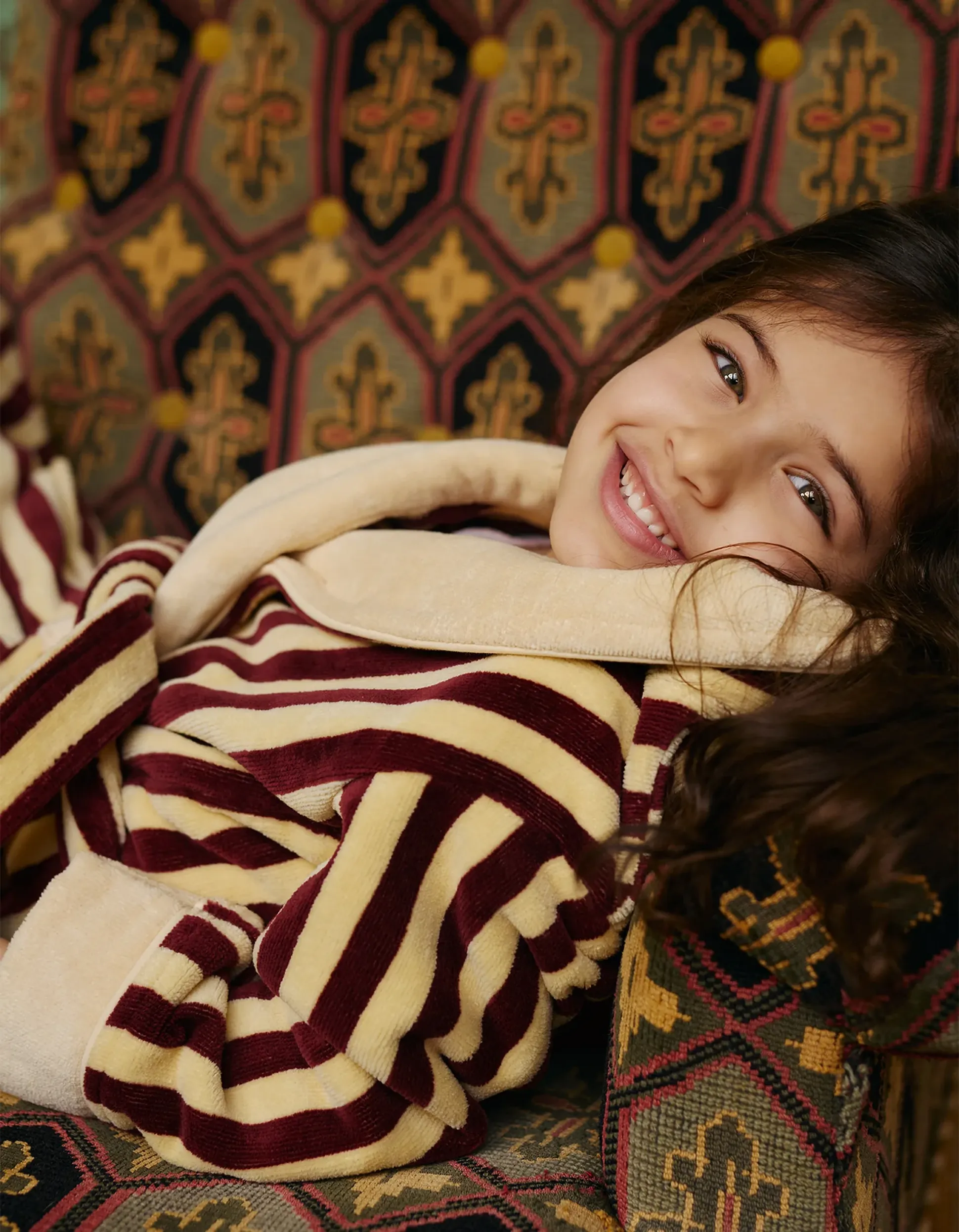 Un enfant souriant, vêtu d'une robe à rayures, est allongé dans un fauteuil à motifs géométriques et floraux.
