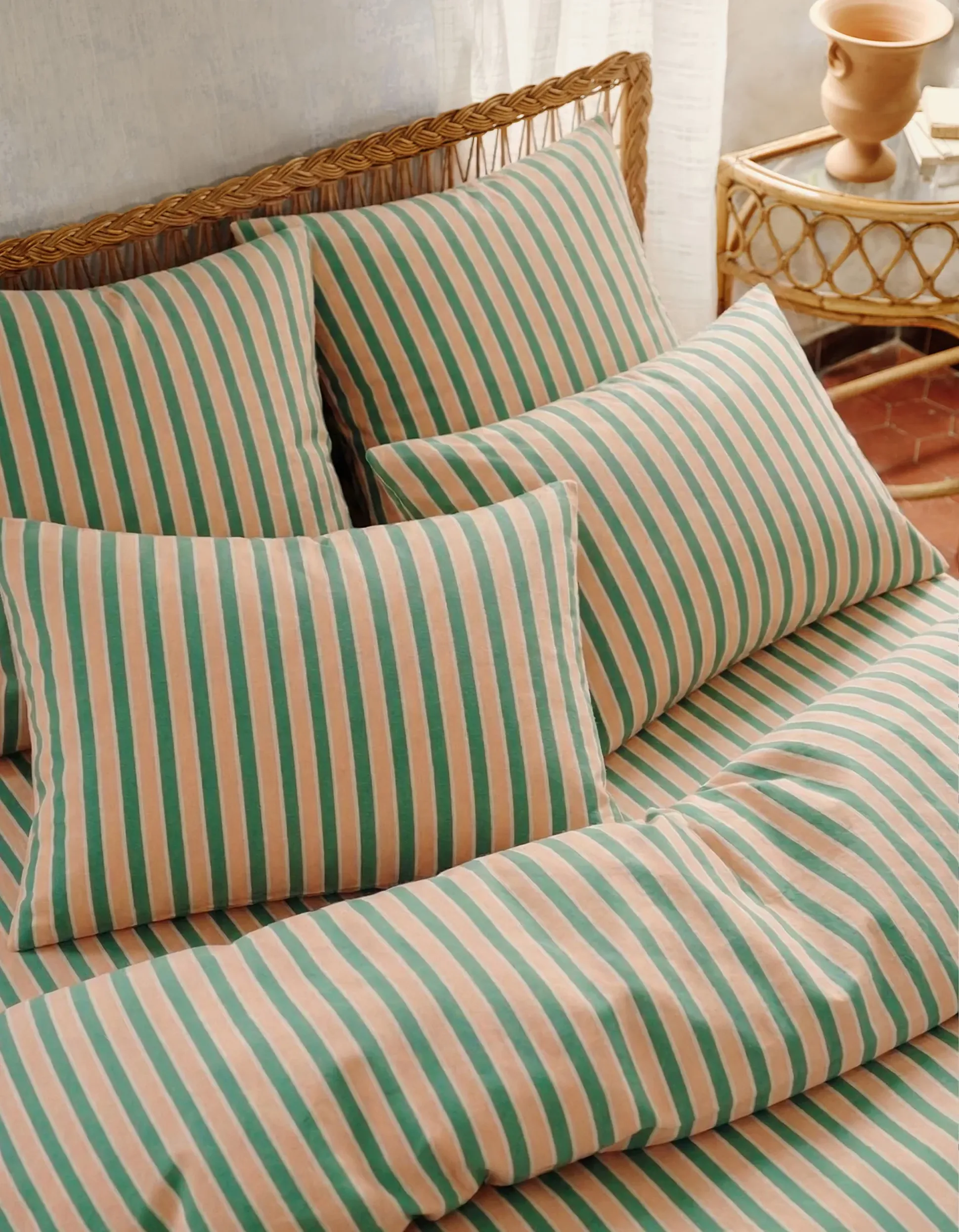 Une parure de lit rayée aux teintes roses et vertes, agrémentée de plusieurs coussins posés sur une tête de lit tissée, à côté d'une petite table d'appoint en osier sur laquelle sont disposés des objets décoratifs.