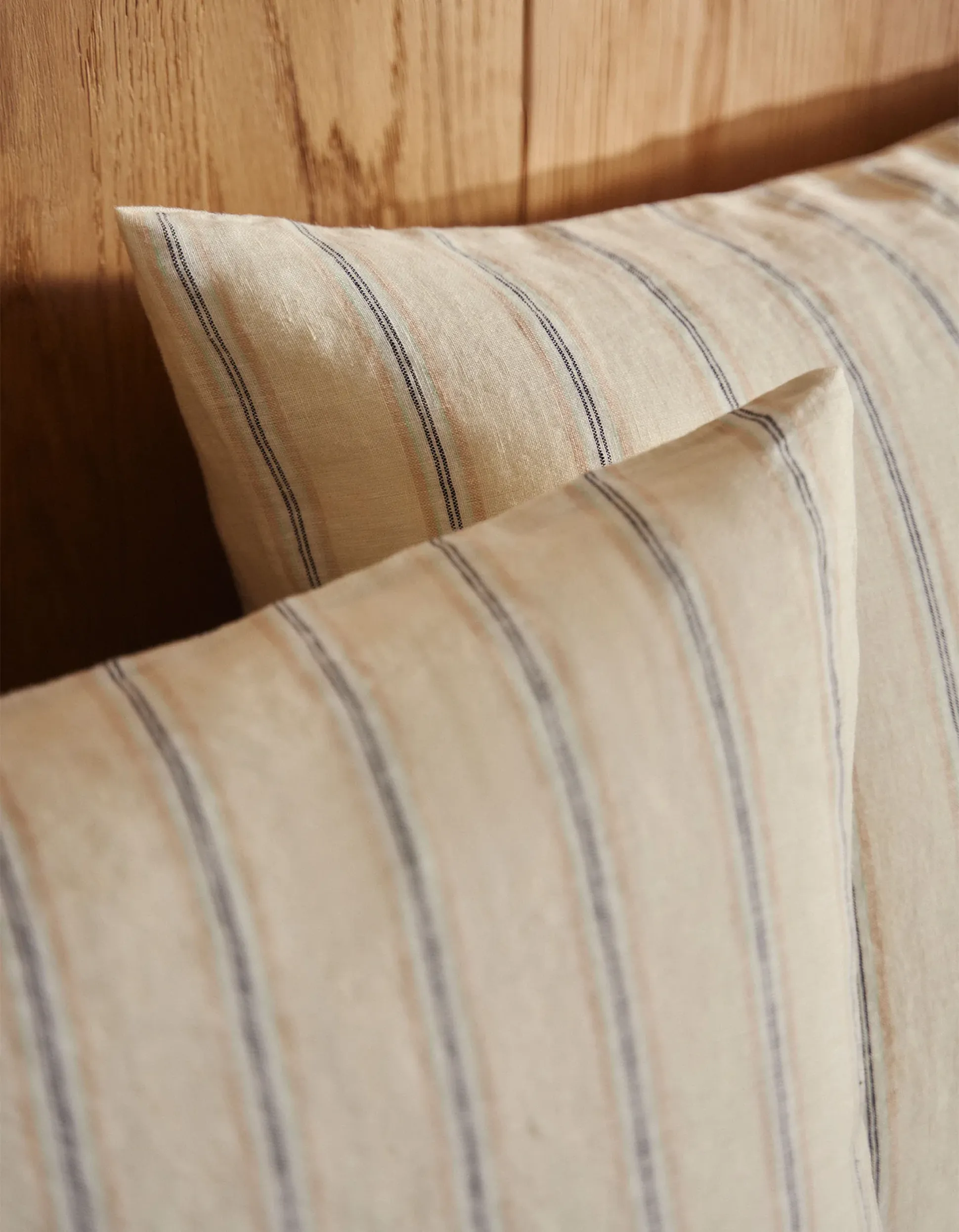 Des coussins beiges à fines rayures bleues, posés contre une tête de lit en bois.