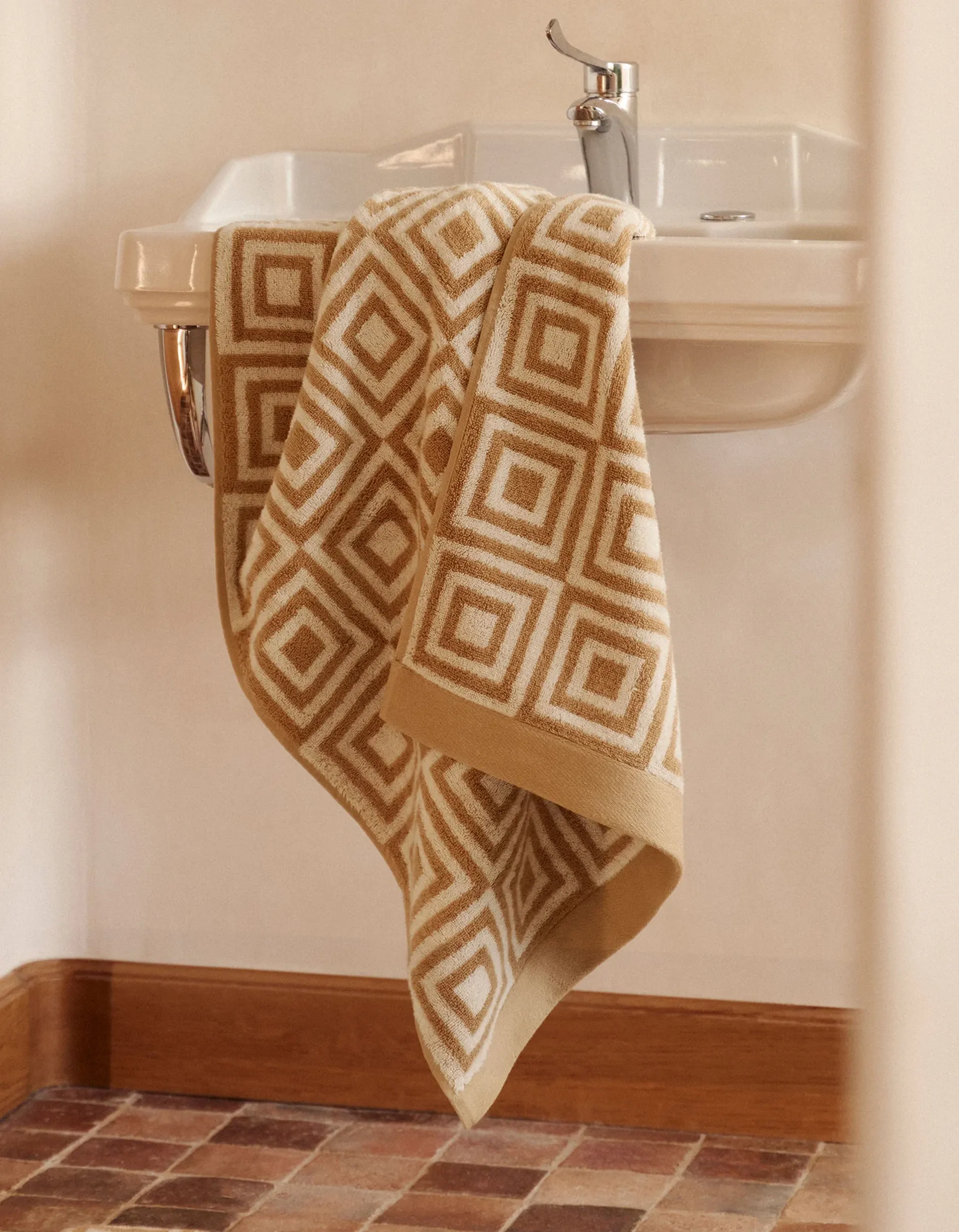 Une serviette beige à motifs géométriques posée sur un lavabo blanc équipé d'un robinet chromé, dans une salle de bains carrelée.
