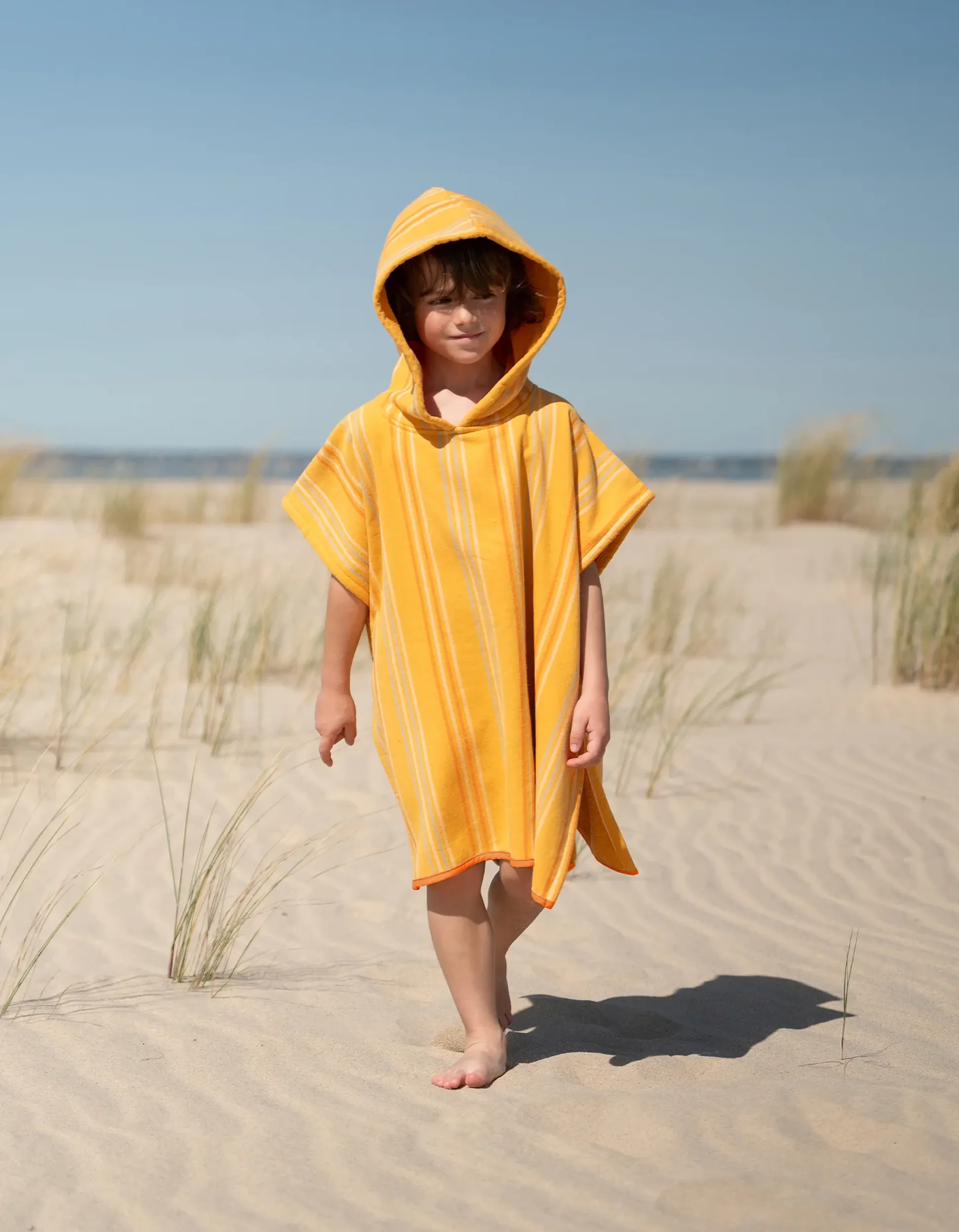Une enfant vêtue d'une robe-serviette jaune à capuche marche pieds nus sur une plage de sable, avec en arrière-plan quelques brins d'herbe et un ciel bleu limpide.