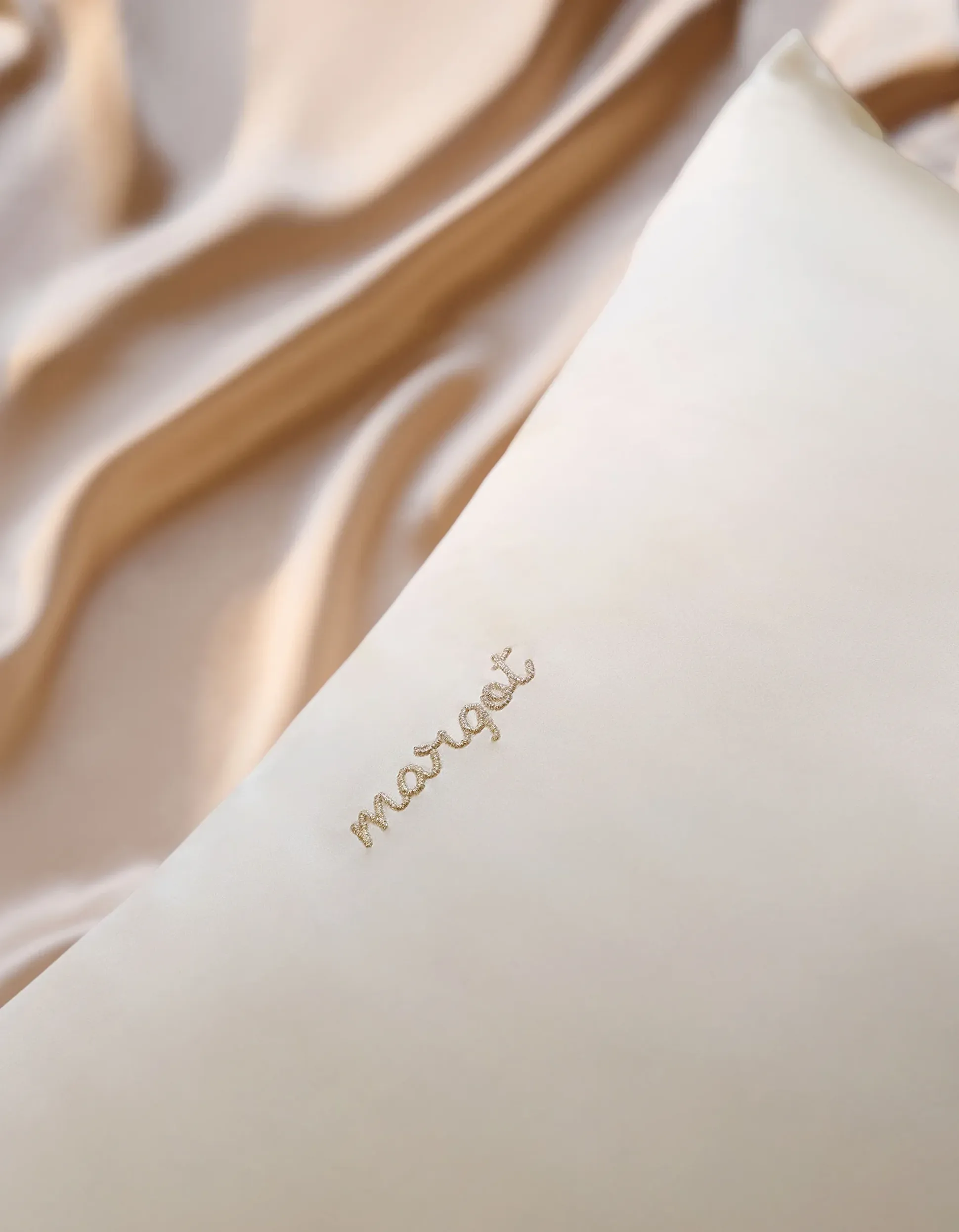 Une taie d'oreiller en soie douce, sur laquelle le prénom « Margot » est brodé en lettres dorées, sur un fond de tissu beige lisse.