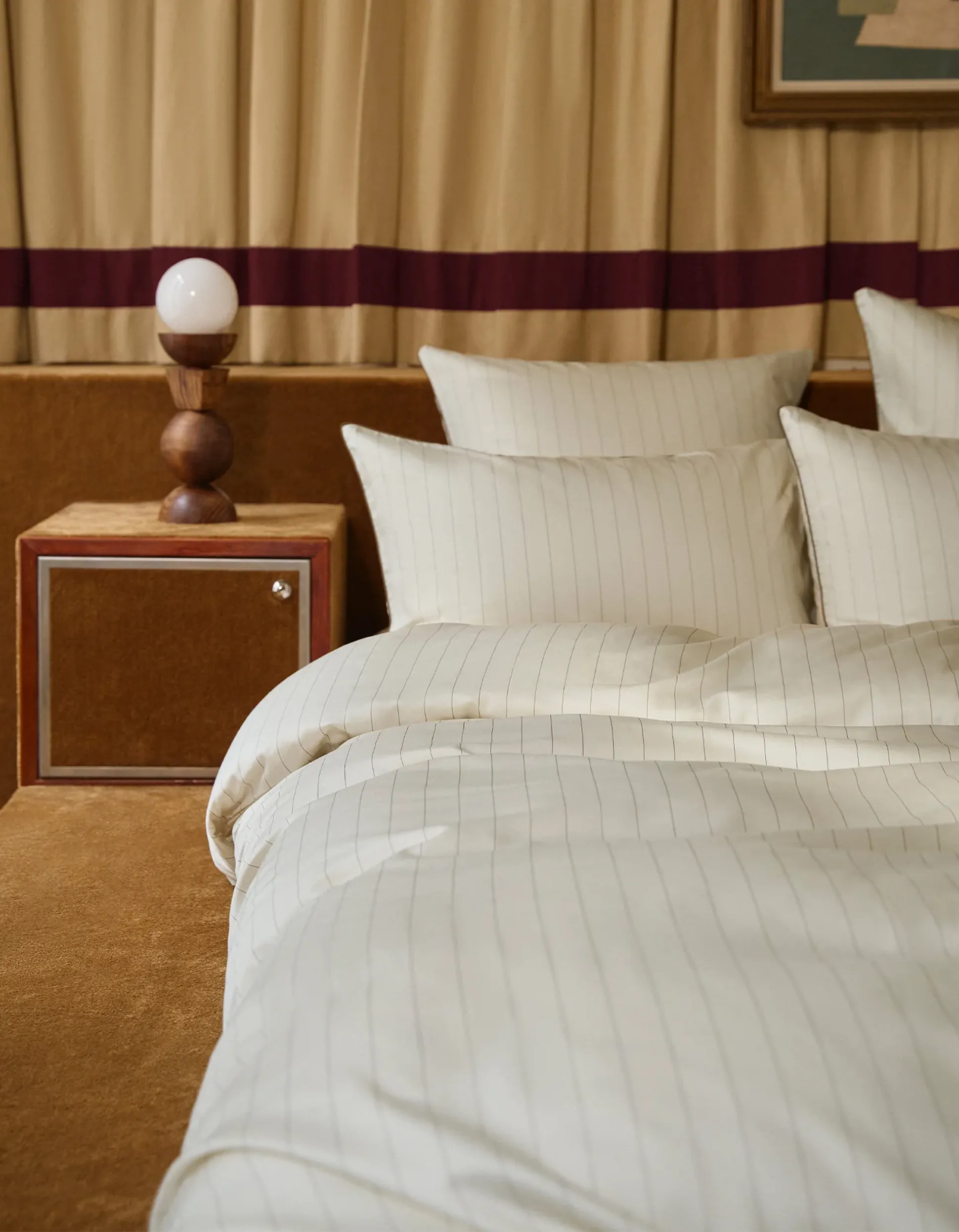 Chambre à coucher avec une parure de lit blanche à rayures, une table de chevet en bois et une lampe originale en bois équipée d'une ampoule blanche ronde. Des rideaux beiges en arrière-plan.