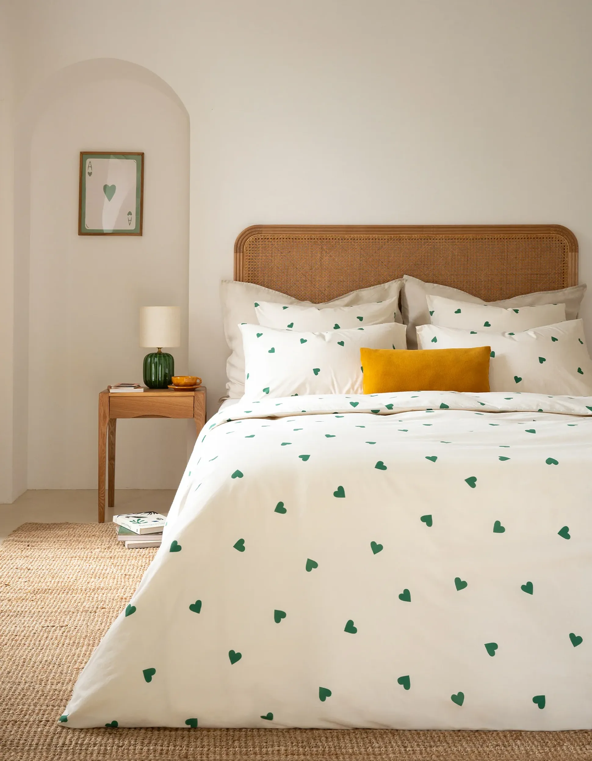 Une chambre douillette avec une tête de lit en rotin, une parure de lit blanche ornée de cœurs verts, un oreiller jaune, une table de chevet en bois et un tableau représentant un cœur accroché au mur.