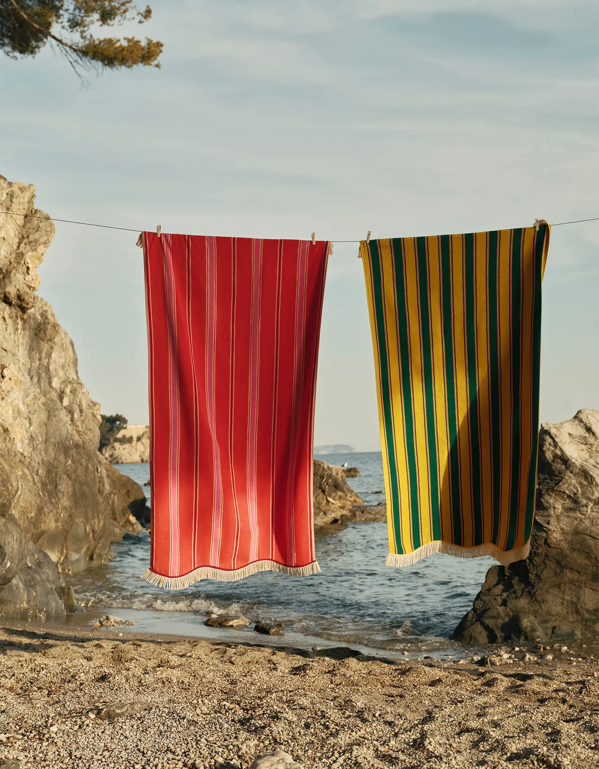 Deux serviettes rayées, l'une rouge et l'autre jaune-vert, sont suspendues à une corde tendue entre des rochers, sur une plage où la mer est calme et le ciel dégagé en arrière-plan.
