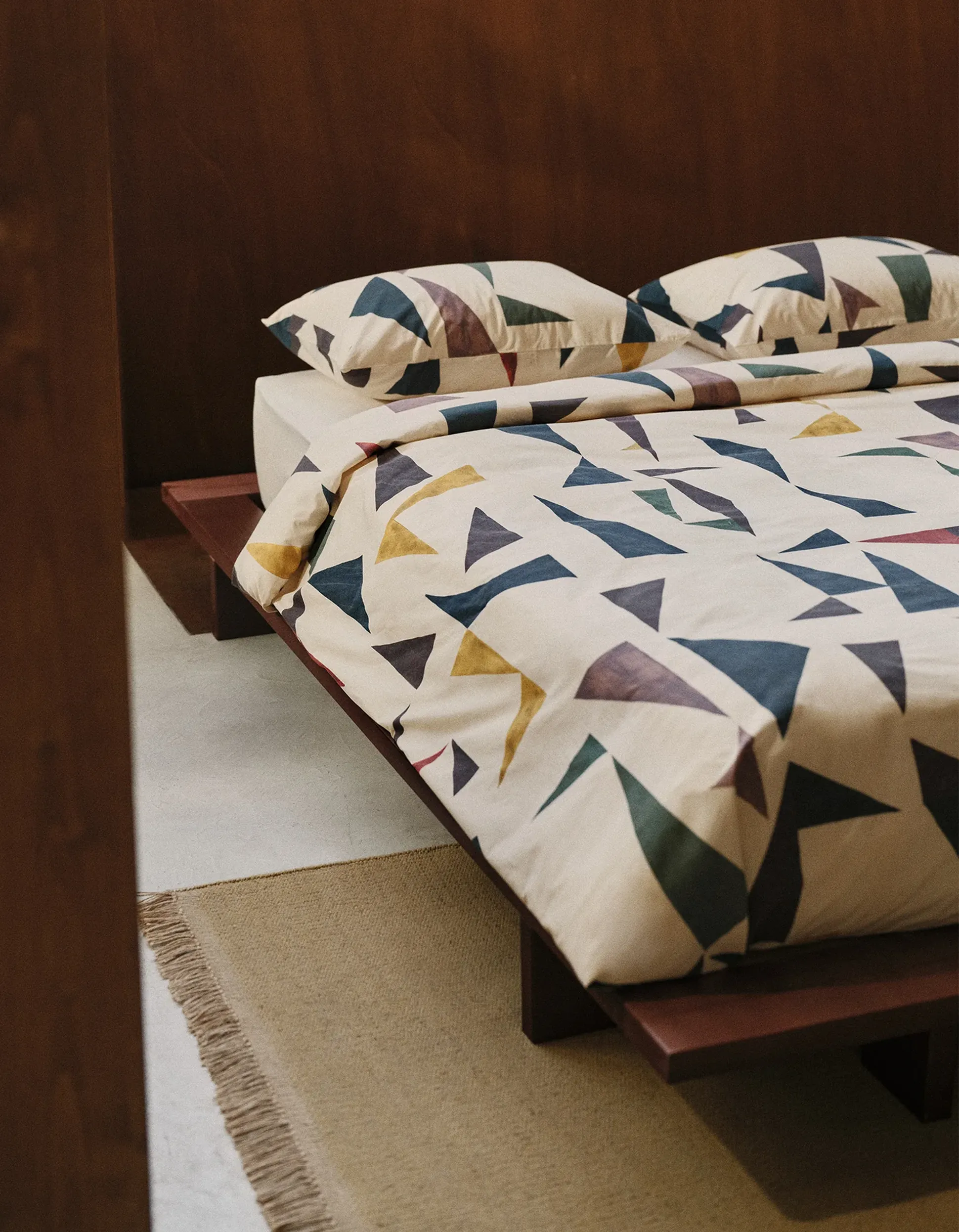 Lit avec couette et oreillers à motifs géométriques, cadre en bois et tapis beige posé sur un sol clair.