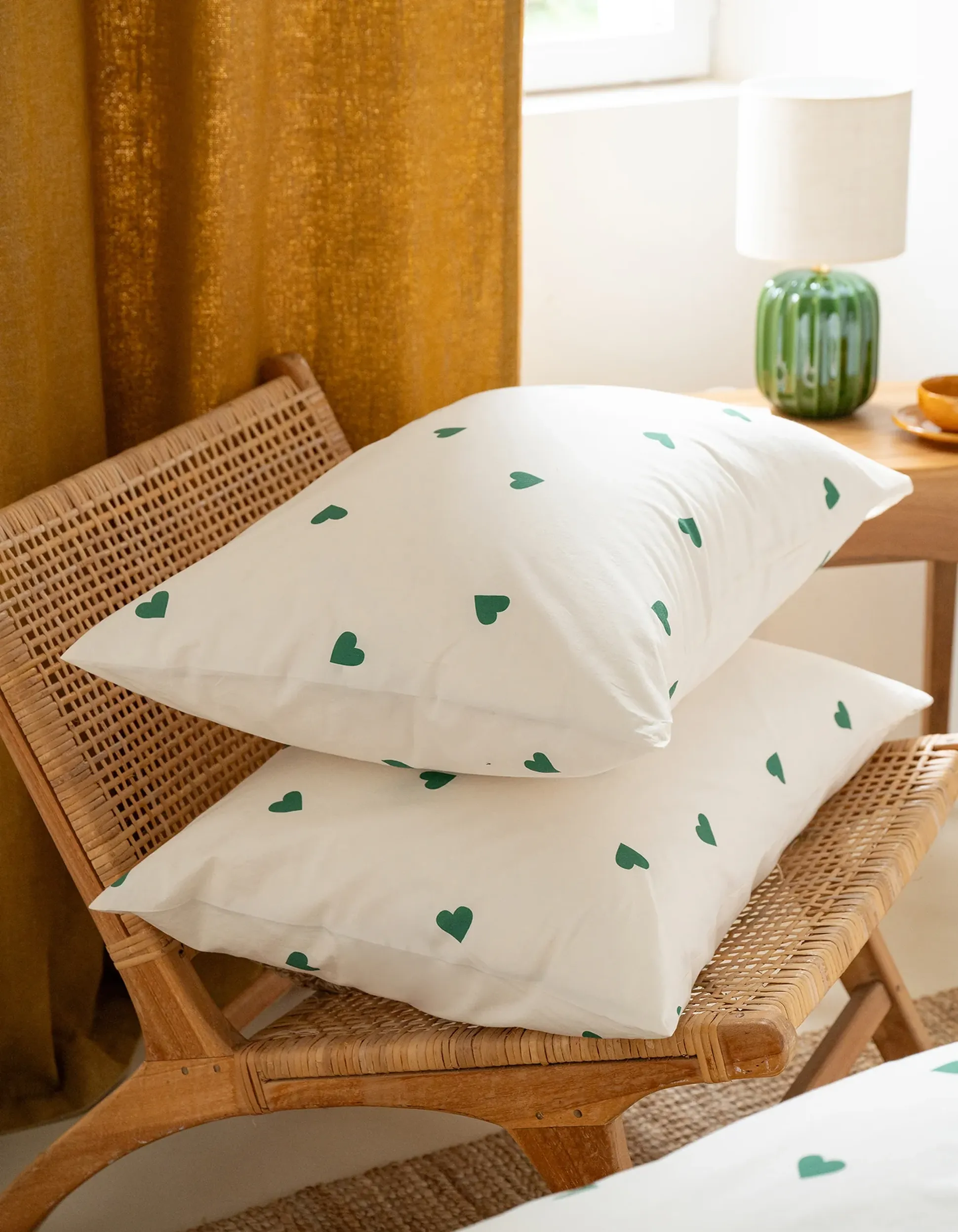Deux coussins blancs ornés de motifs de cœurs verts sur une chaise en osier, à côté d'une table sur laquelle se trouve une lampe verte, avec en arrière-plan des rideaux couleur moutarde.