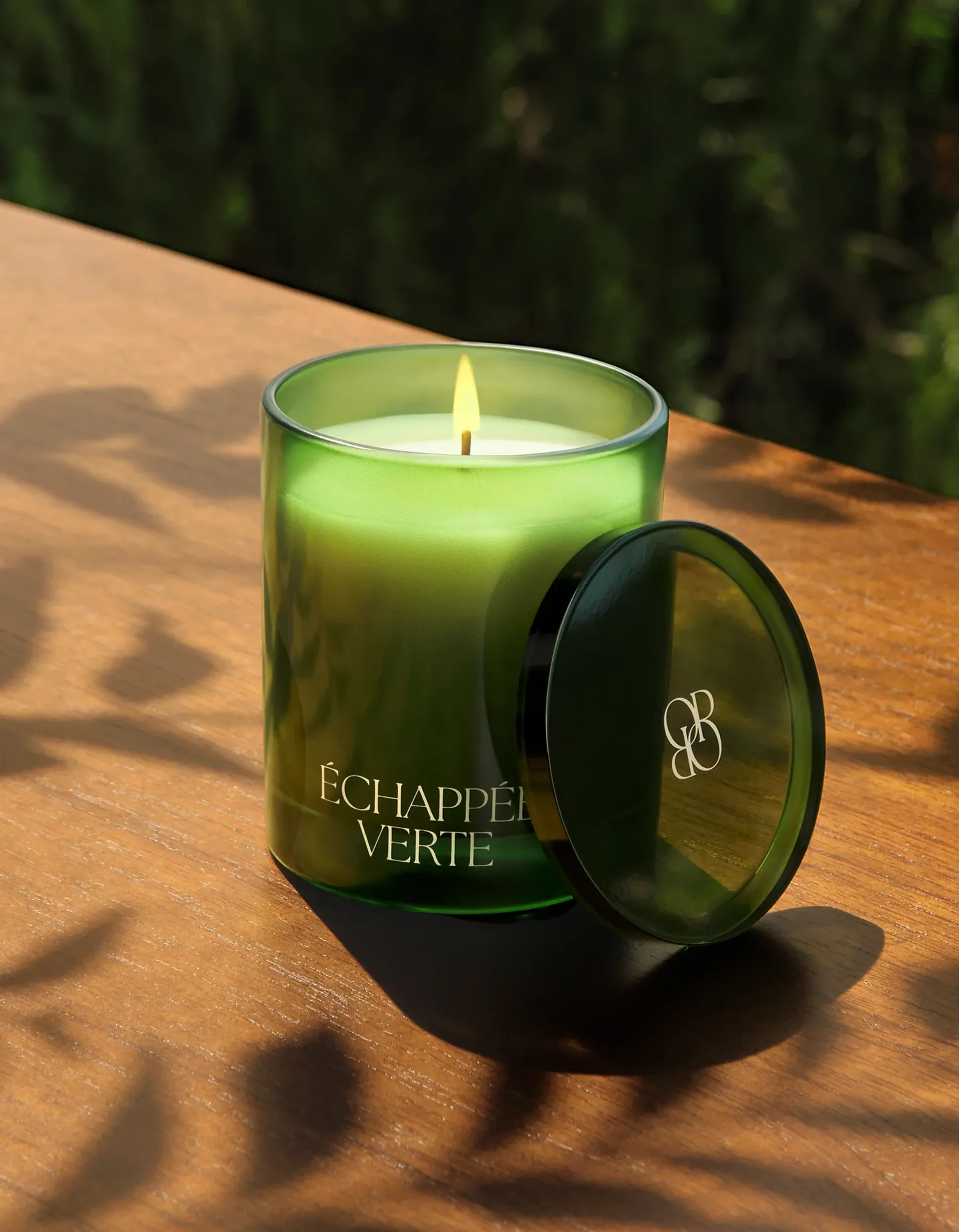 Bougie verte étiquetée « Échappée Verte » sur une surface en bois, couvercle entrouvert, avec des ombres de feuillage.