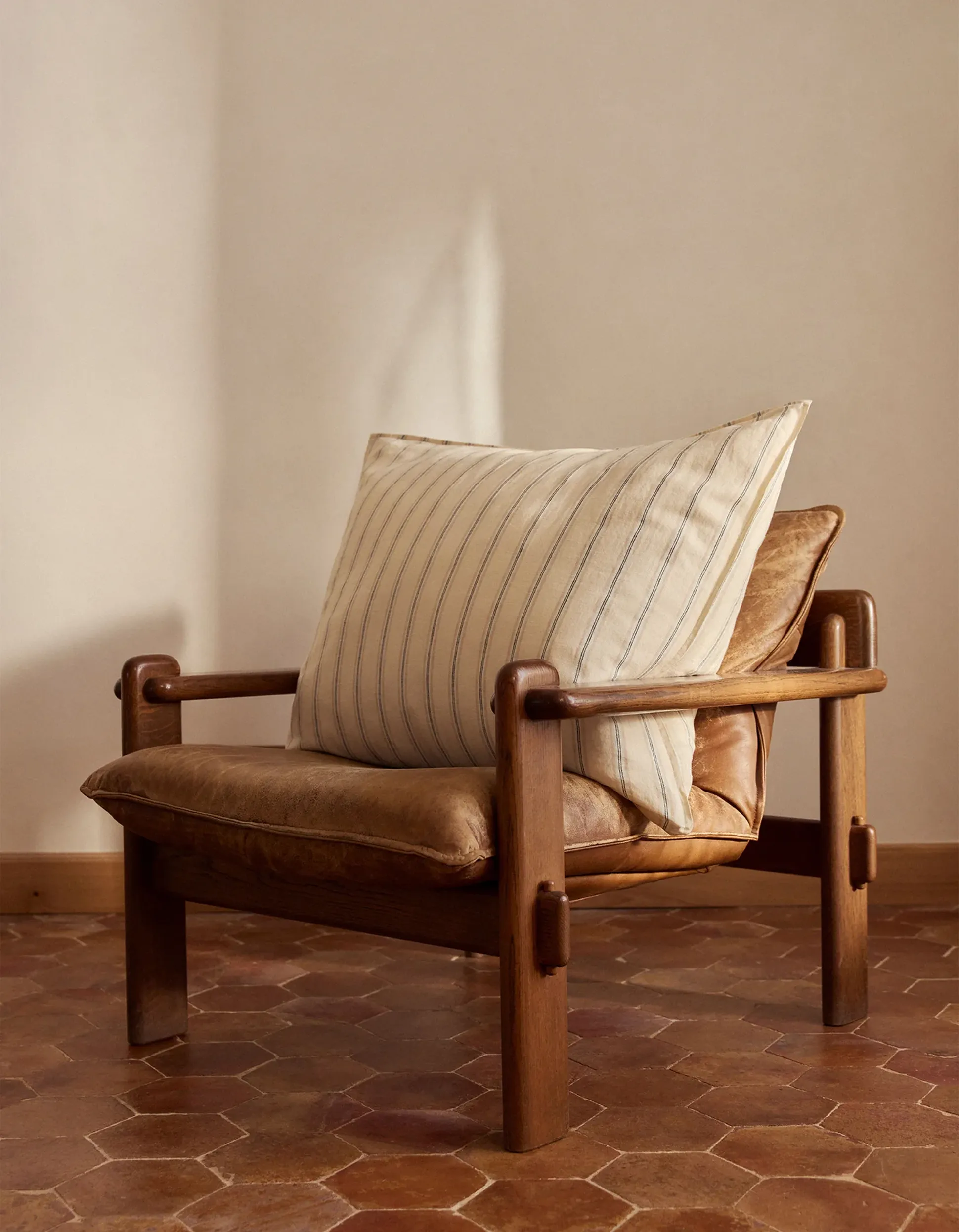 Une chaise en bois rembourrée et revêtue de cuir est placée sur un sol en carreaux hexagonaux ; elle est agrémentée d'un coussin rayé et disposée contre un mur aux tons neutres.