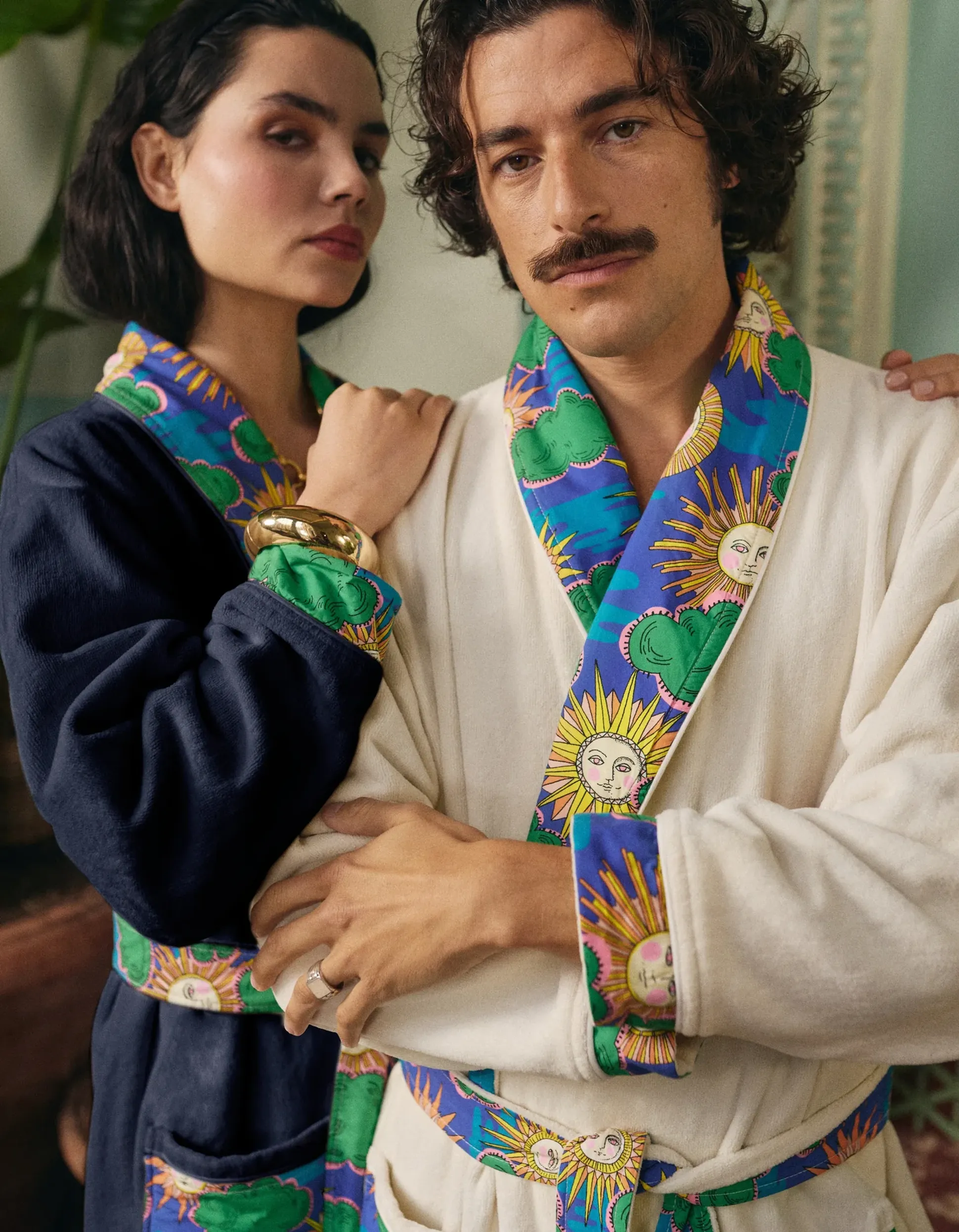 Un homme et une femme posent ensemble, vêtus de robes à motifs représentant des soleils et des nuages, debout à l'intérieur, le visage empreint d'une expression discrète.