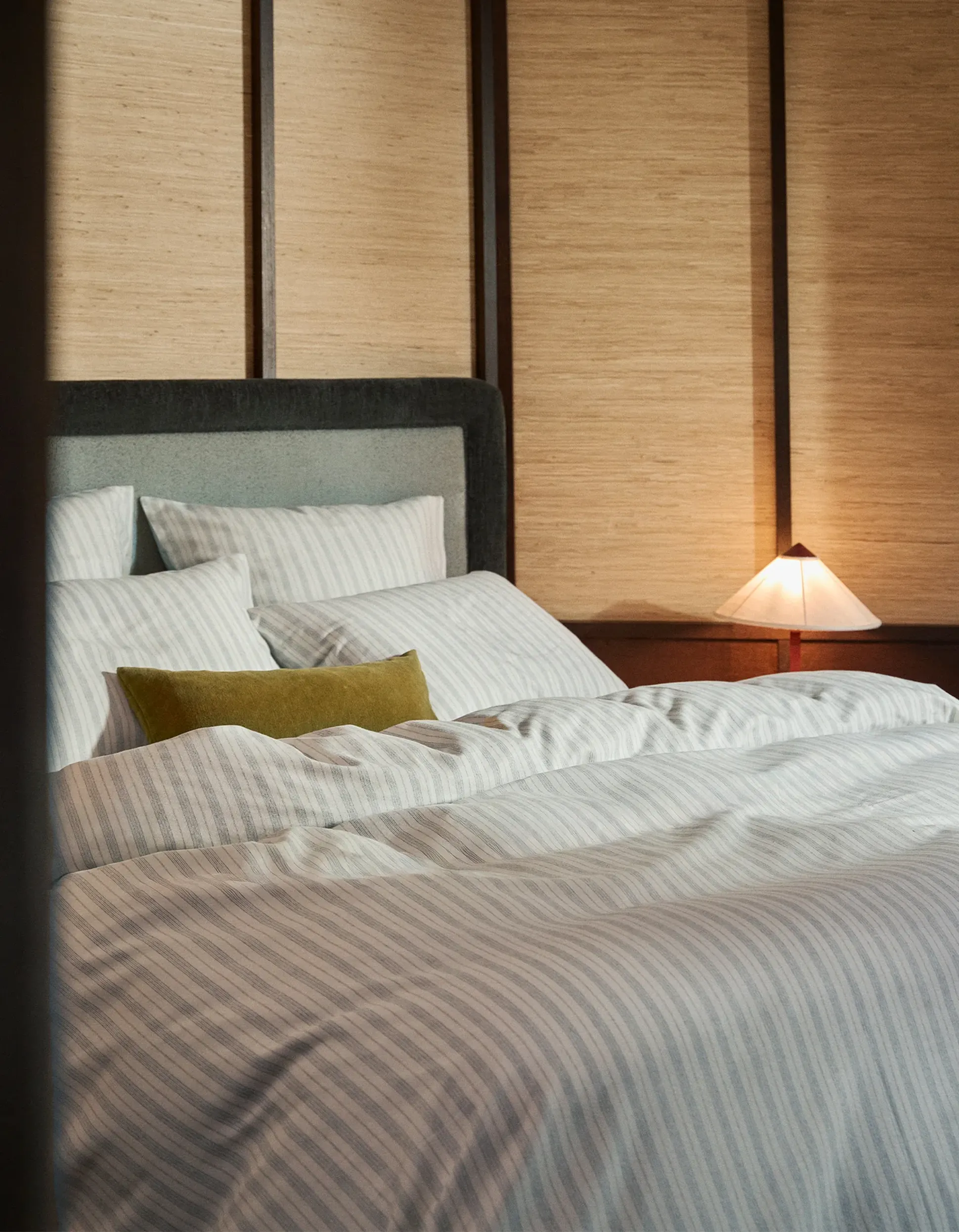 Une chambre douillette avec un couvre-lit rayé, un coussin décoratif vert et une lampe de chevet aux lueurs chaleureuses, le tout devant un mur lambrissé.
