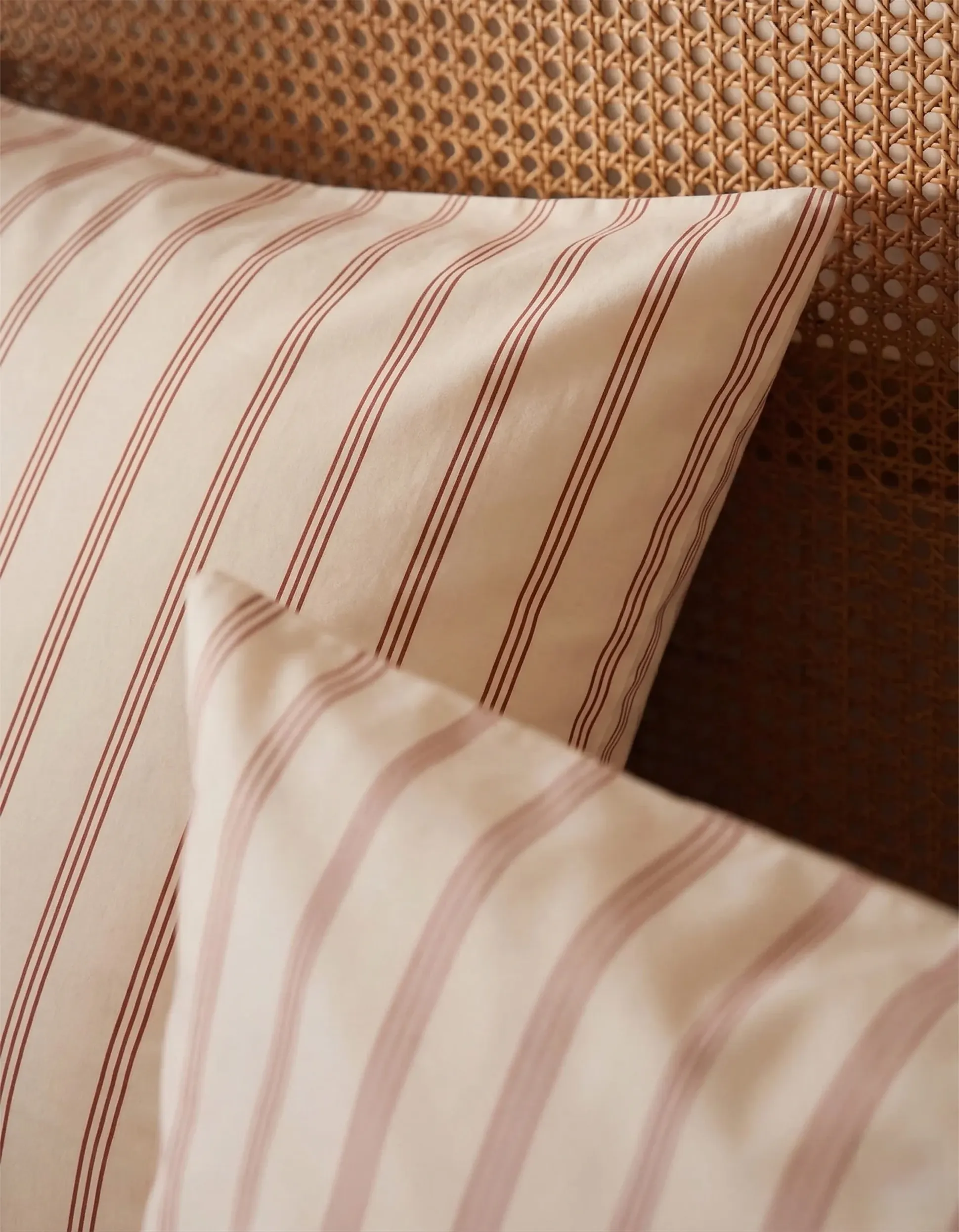Deux coussins couleur crème à fines rayures rouges appuyés contre une tête de lit en osier.