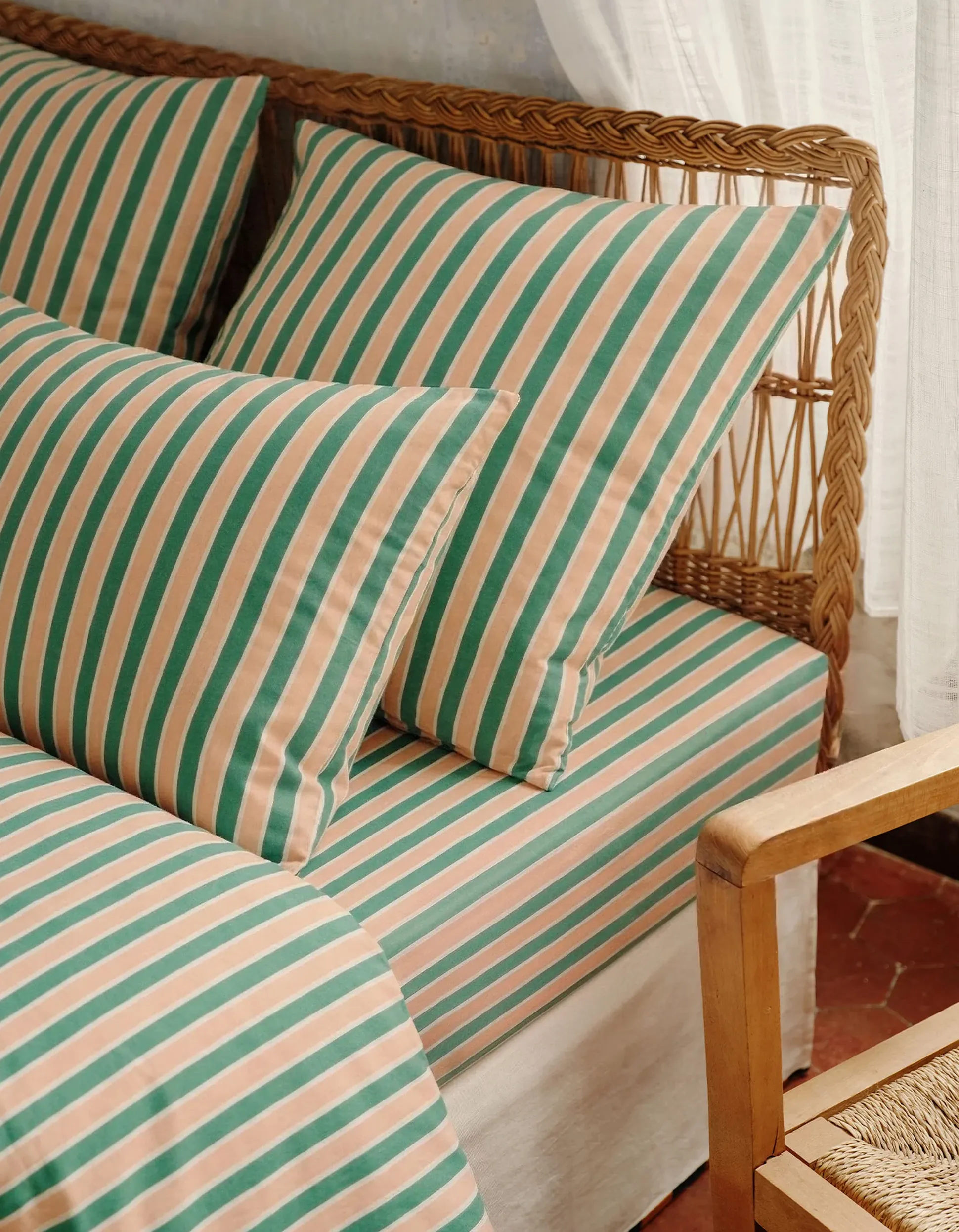 Un lit douillet, avec des oreillers et des draps rayés de vert et de beige, adossé à une tête de lit en osier, à côté d'une chaise en bois et de rideaux voilés.